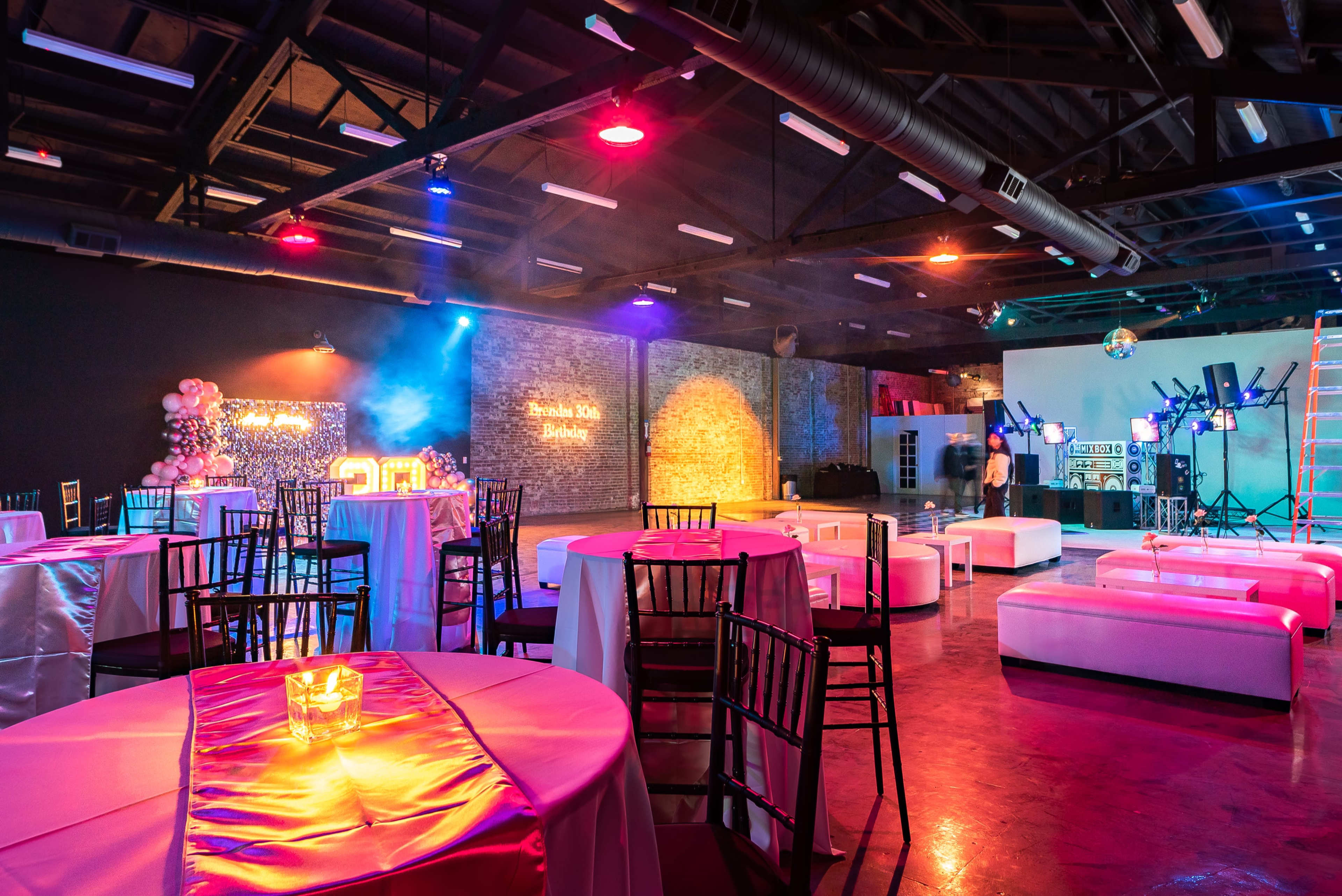 The image shows a spacious event venue with decorated tables, a lounge area, and colorful lighting set up for a celebration.