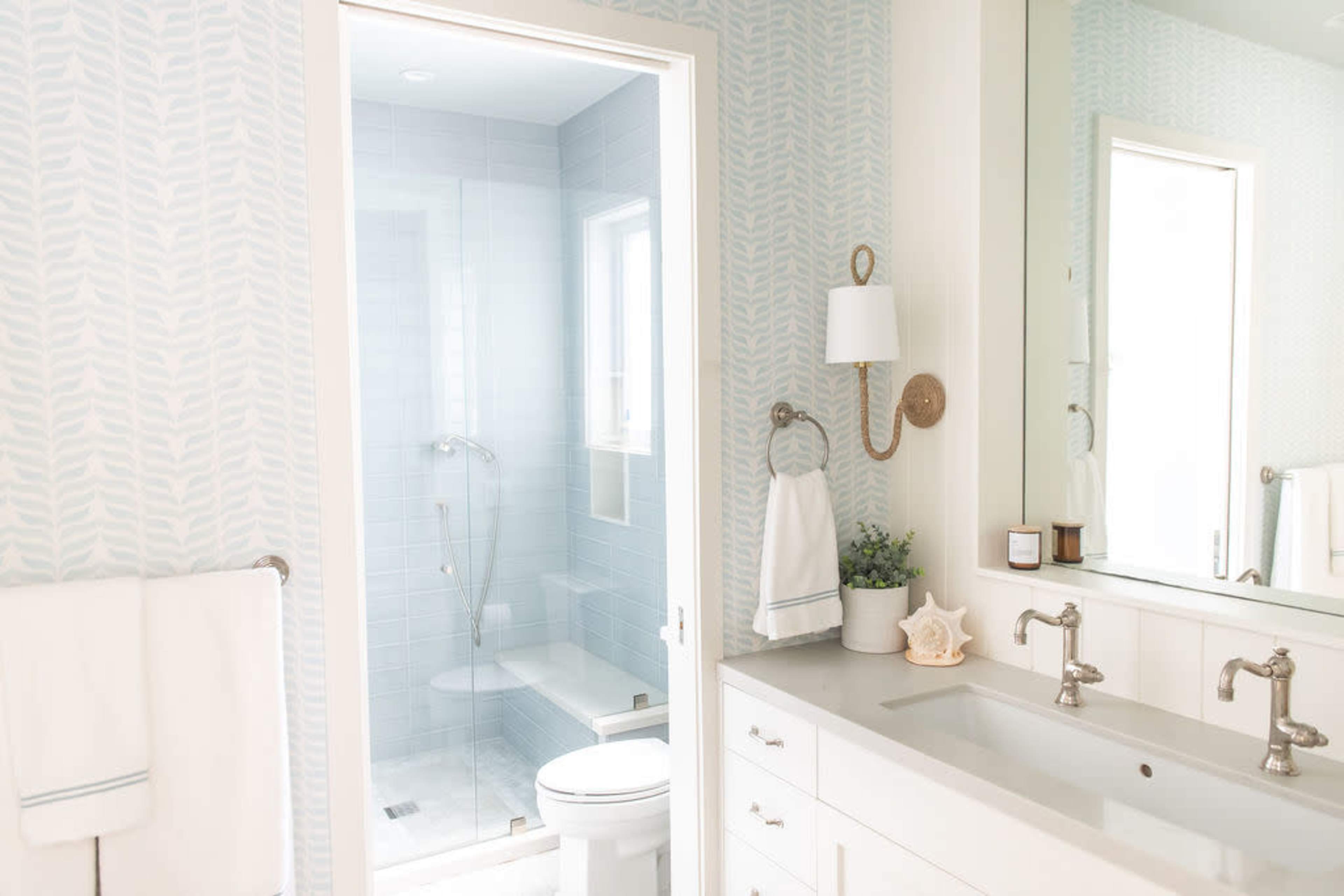 The image shows a modern bathroom featuring a walk-in shower, a double vanity with sinks, and light blue patterned wallpaper.