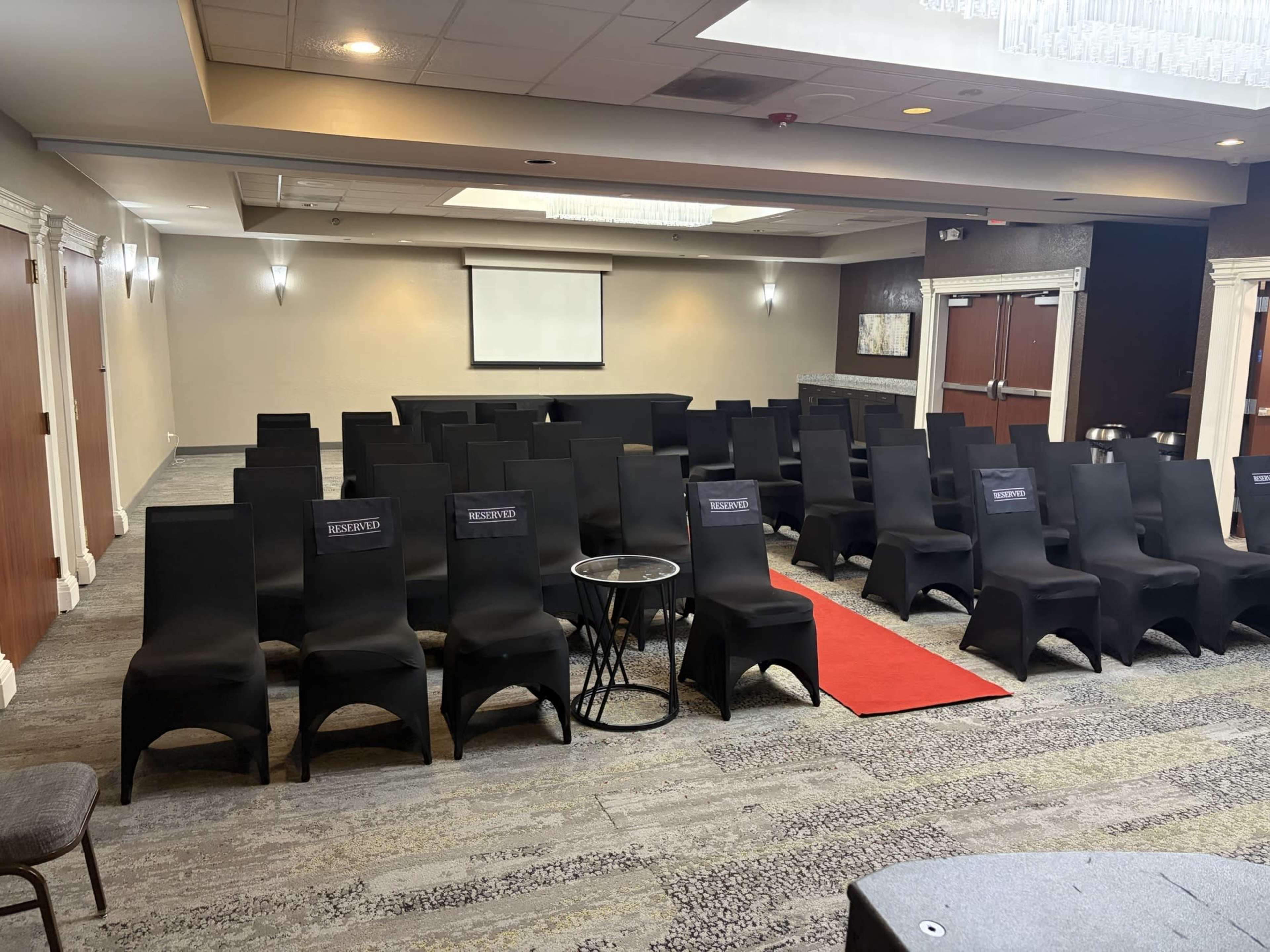 A conference room is set up with rows of black chairs and a red carpet leading to a stage, with a projector screen at the front.