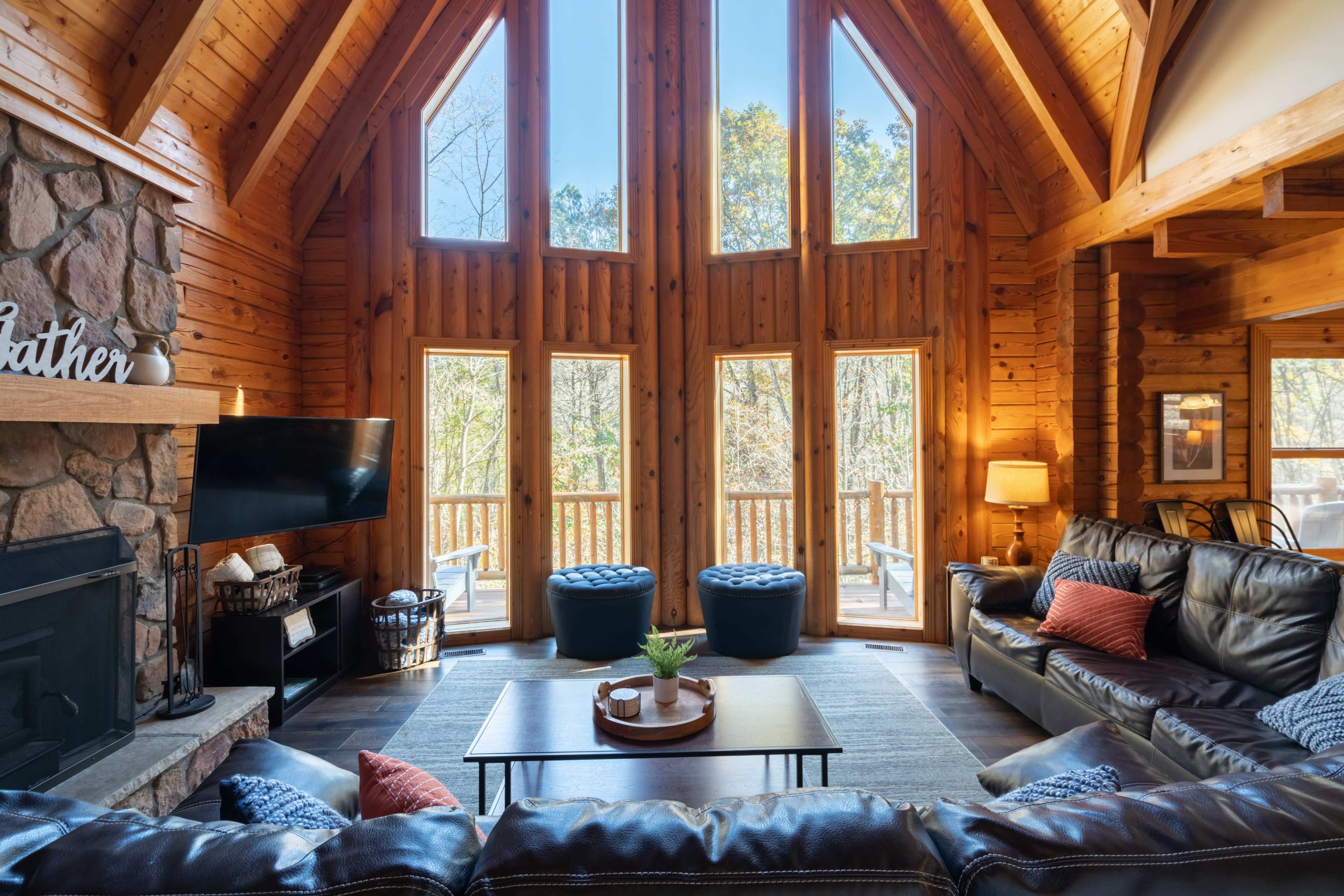 The living room features large windows that frame a view of trees, wooden walls, a stone fireplace, and leather seating arrangements.