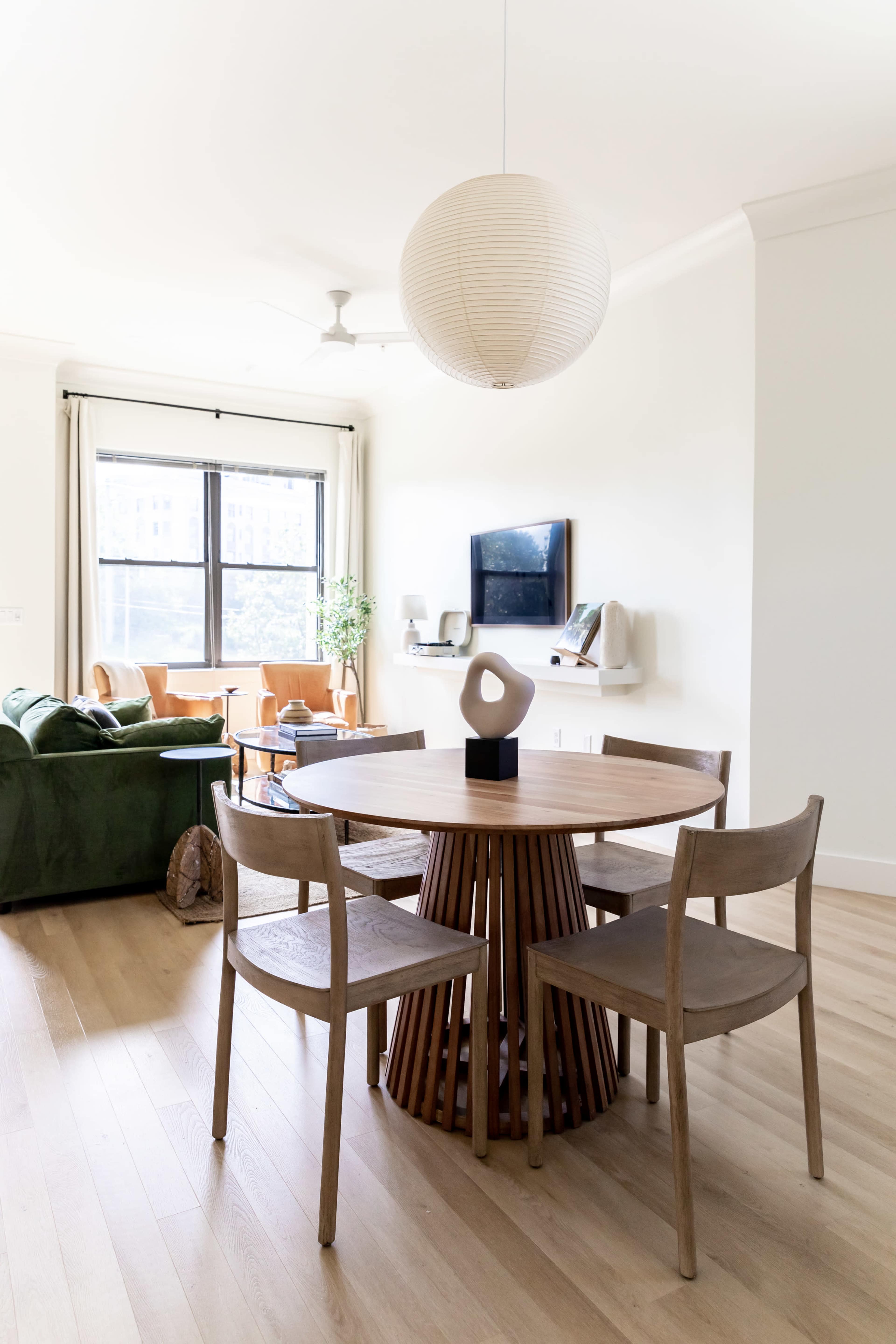 A round wooden dining table with four chairs is positioned in a bright room that features a green sofa and a television mounted on the wall.
