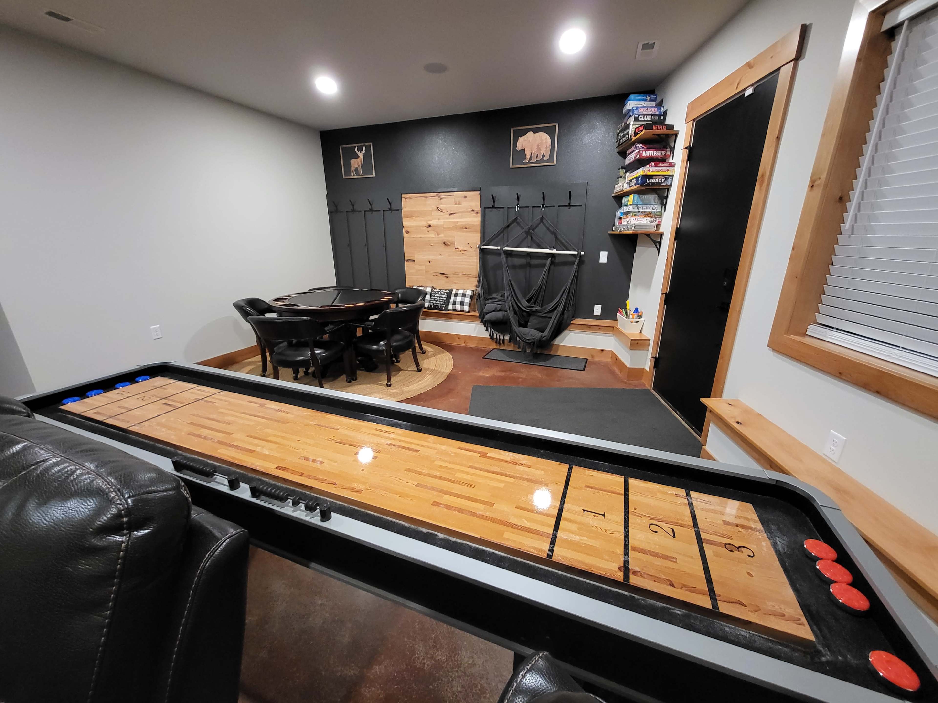 The image shows a game room featuring a shuffleboard table on the left, a round poker table in the back, and shelves filled with board games on the right.