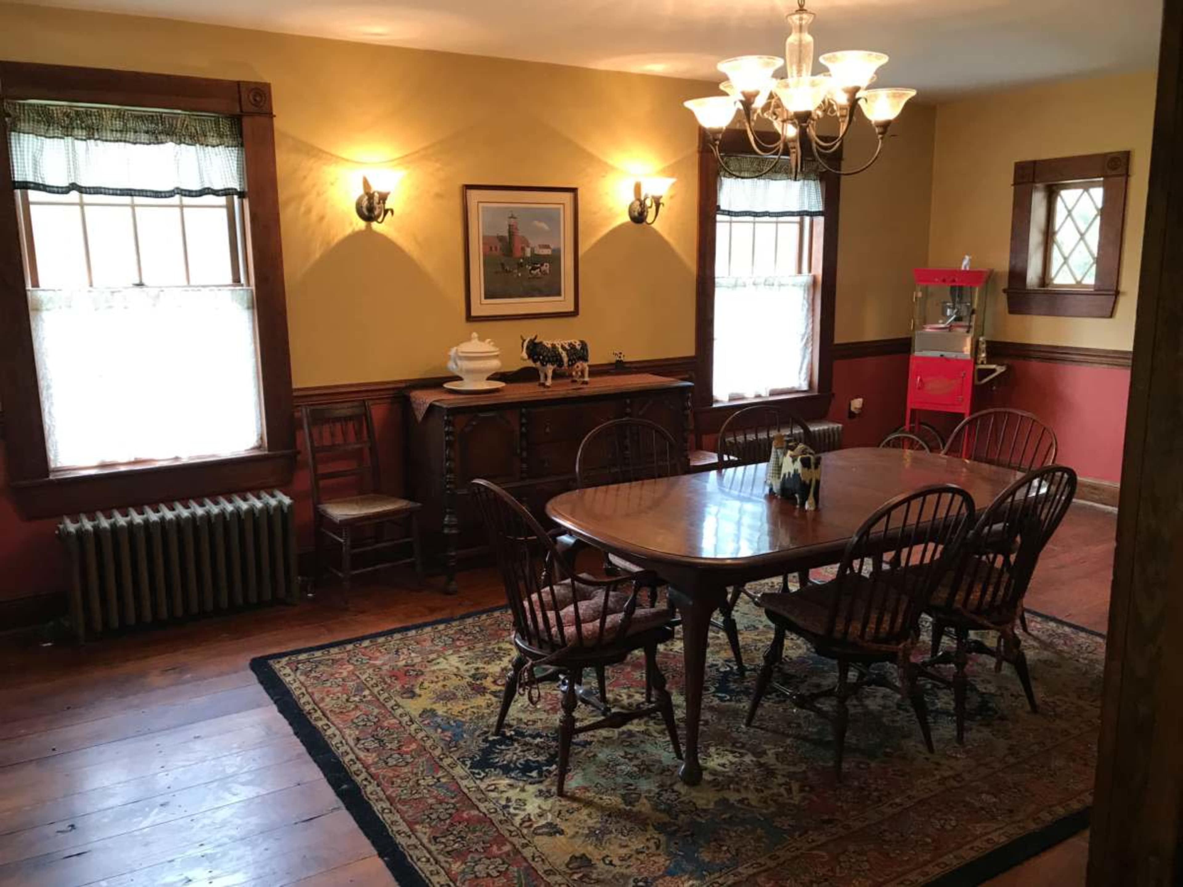 The image shows a dining room with a wooden table surrounded by chairs, a sideboard, and a popcorn machine in one corner, illuminated by a chandelier and wall sconces.