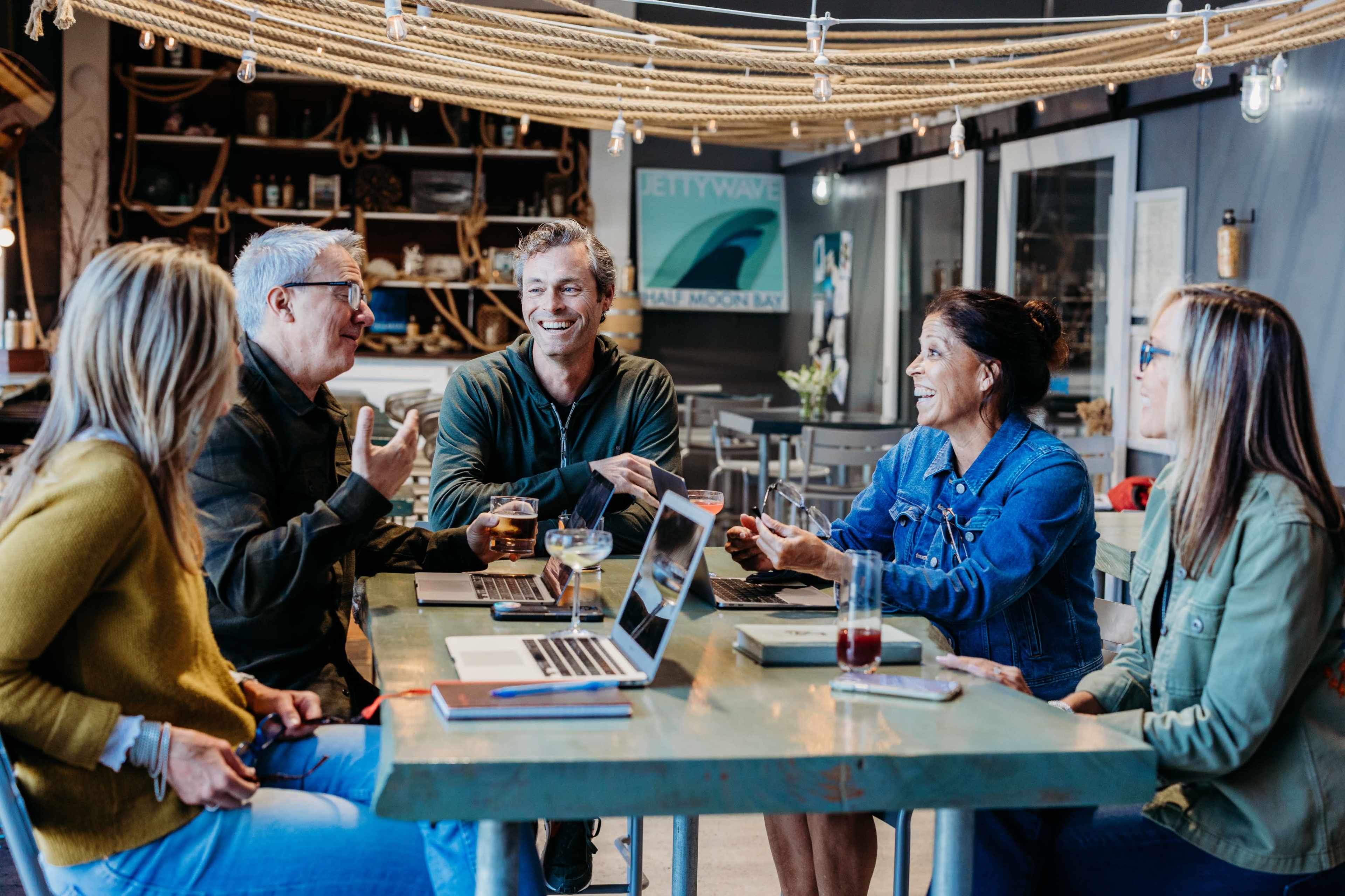 A group of six people engages in a discussion around a table with laptops and drinks in a cozy, well-lit café.