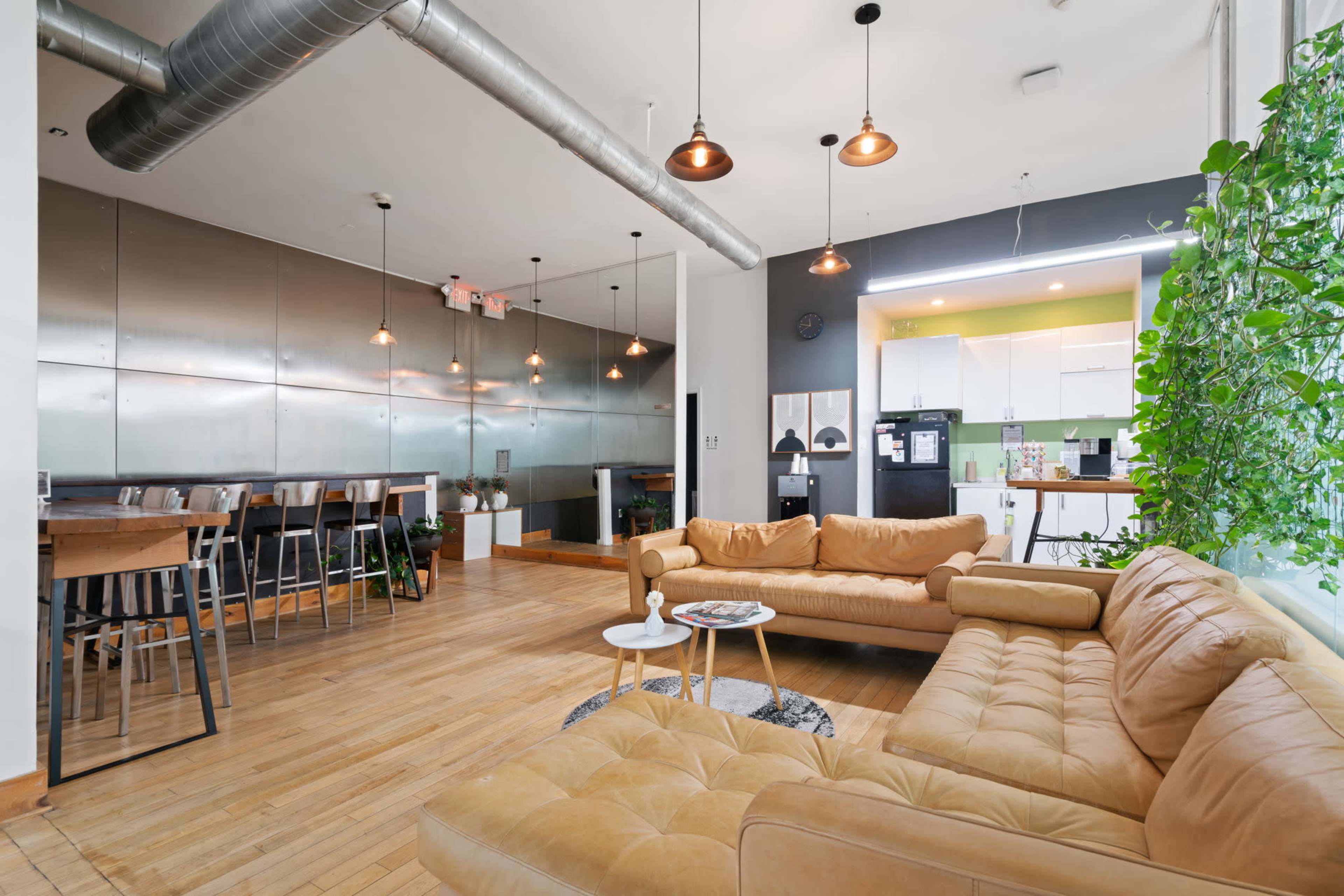 The image shows a modern living space featuring a tan sectional sofa, a dining area with high stools, and a kitchen with stainless steel appliances.