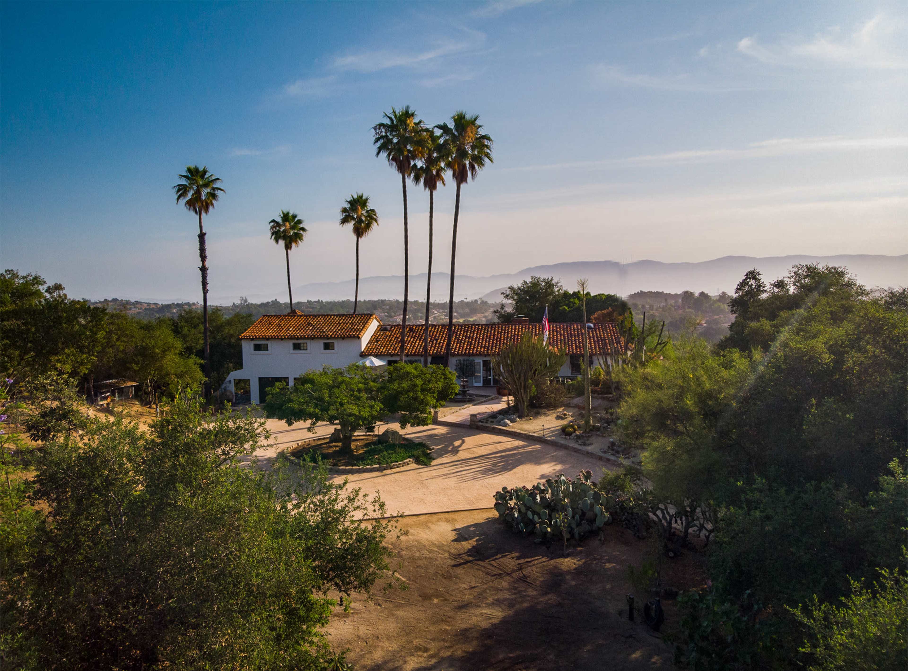 A white house with a red tile roof surrounded by palm trees and scenic hills in the background.