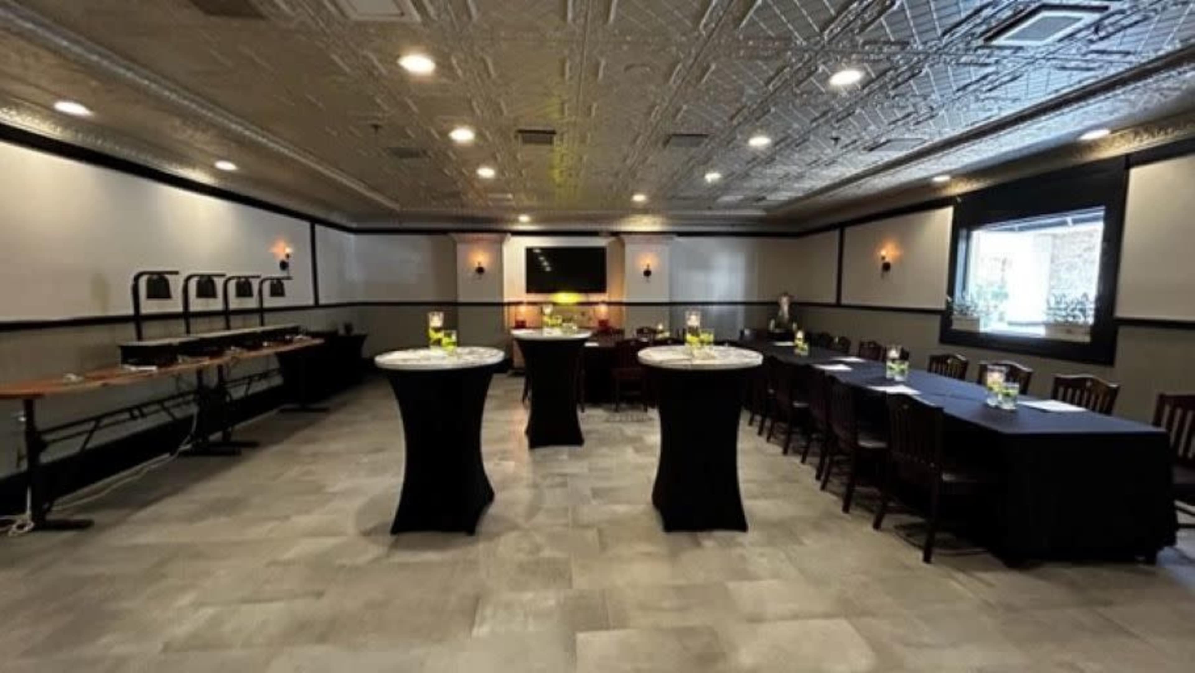 An empty dining room set up for an event, featuring several round tables with black covers, a long table along one wall, and a light-colored ceiling with decorative tiles.