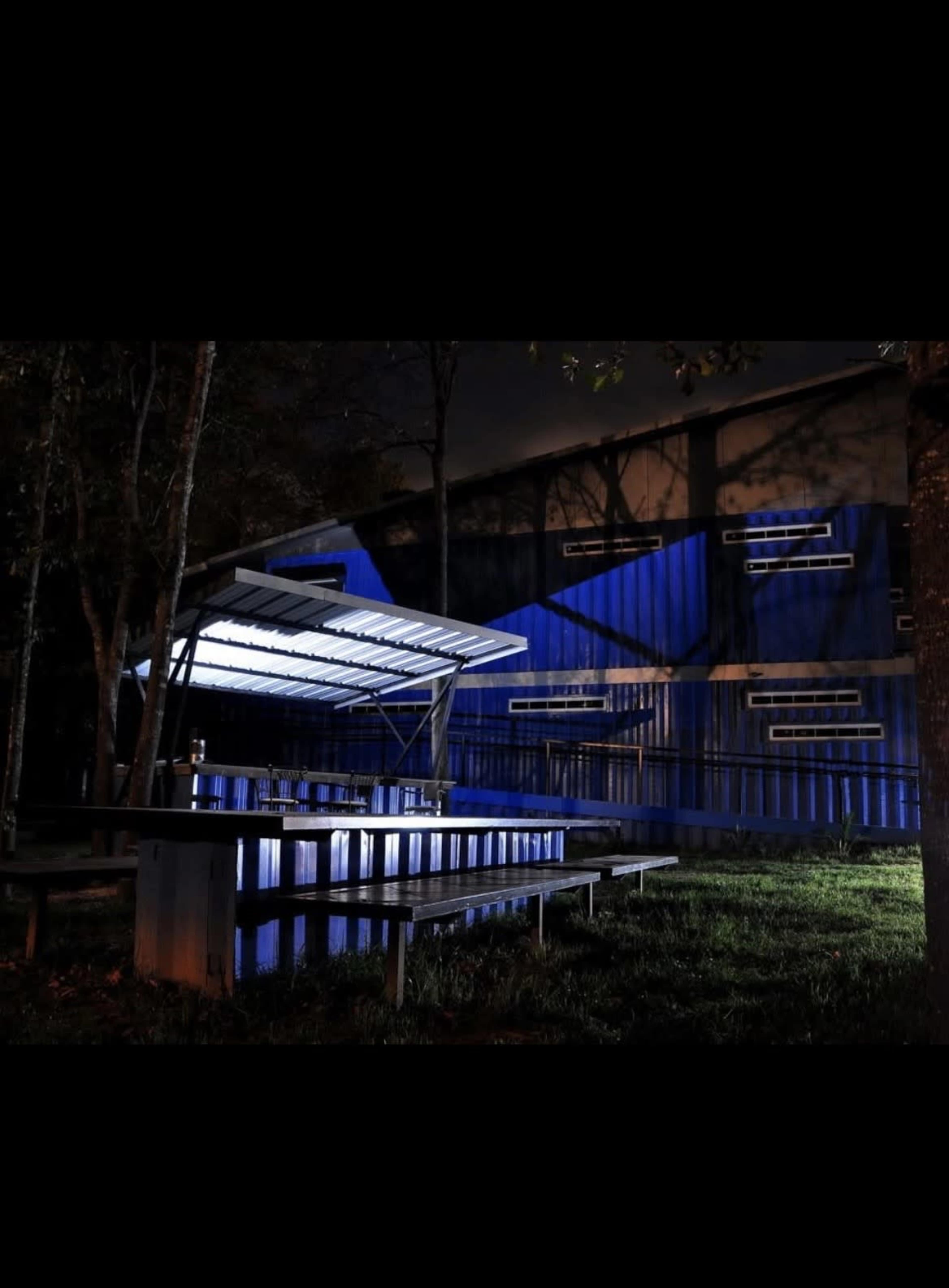 A modern outdoor seating area with a shaded table and illuminated elements, set against a backdrop of a blue container-like structure and trees at night.