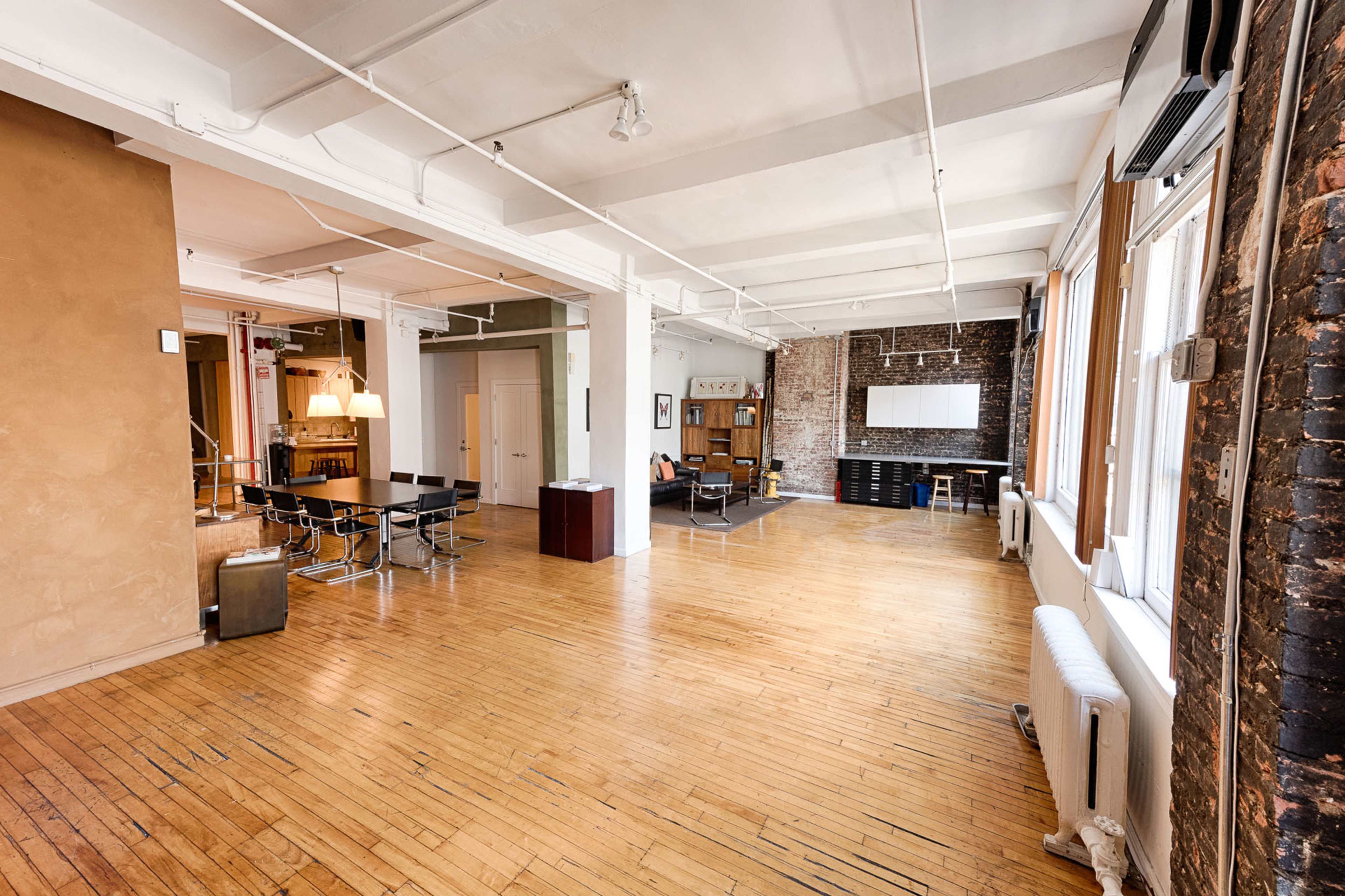 A spacious, open loft interior with hardwood floors, exposed brick walls, and large windows.