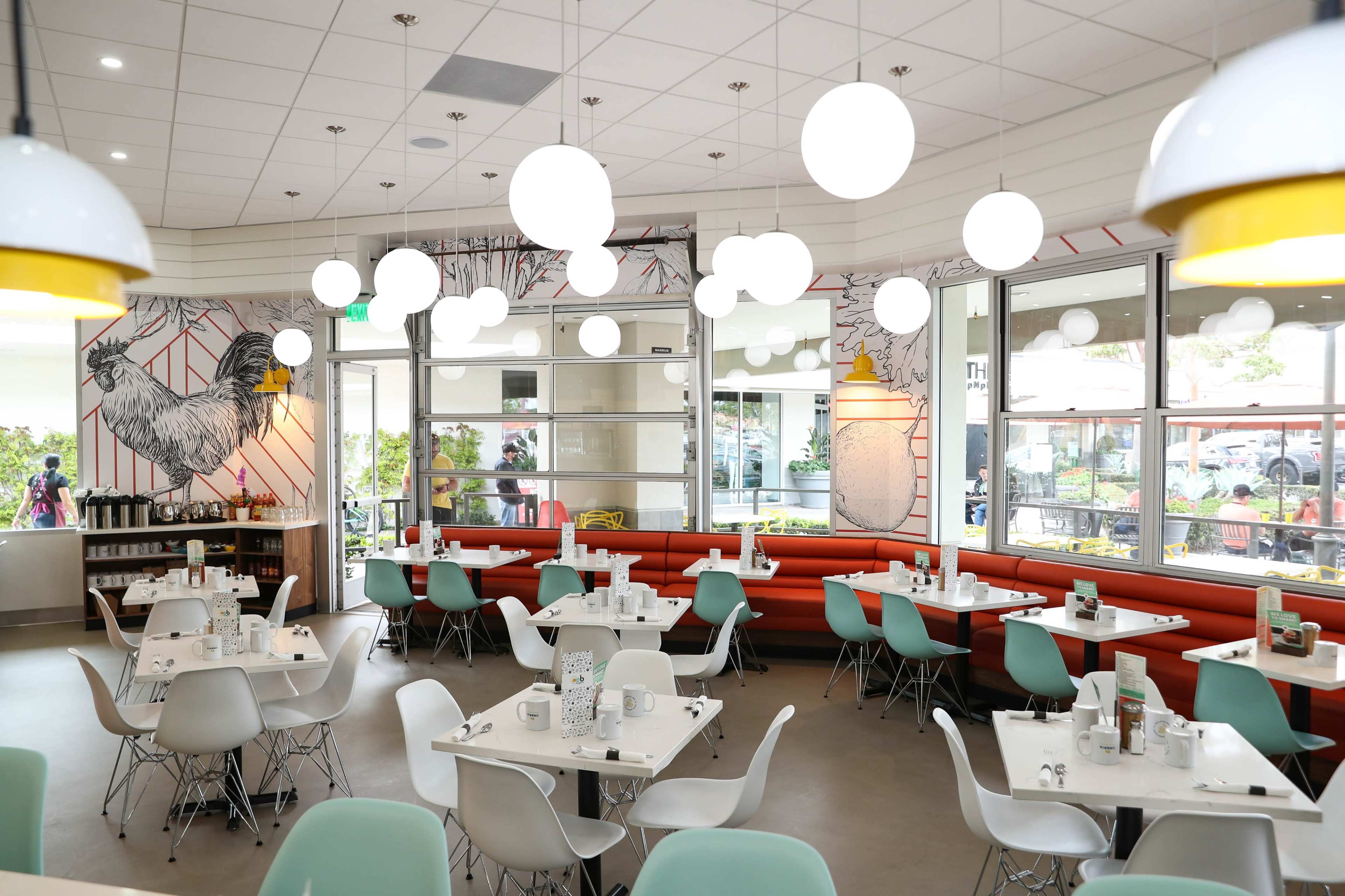 A brightly lit restaurant interior featuring white tables, Eames-style chairs, and large round pendant lights suspended from the ceiling.