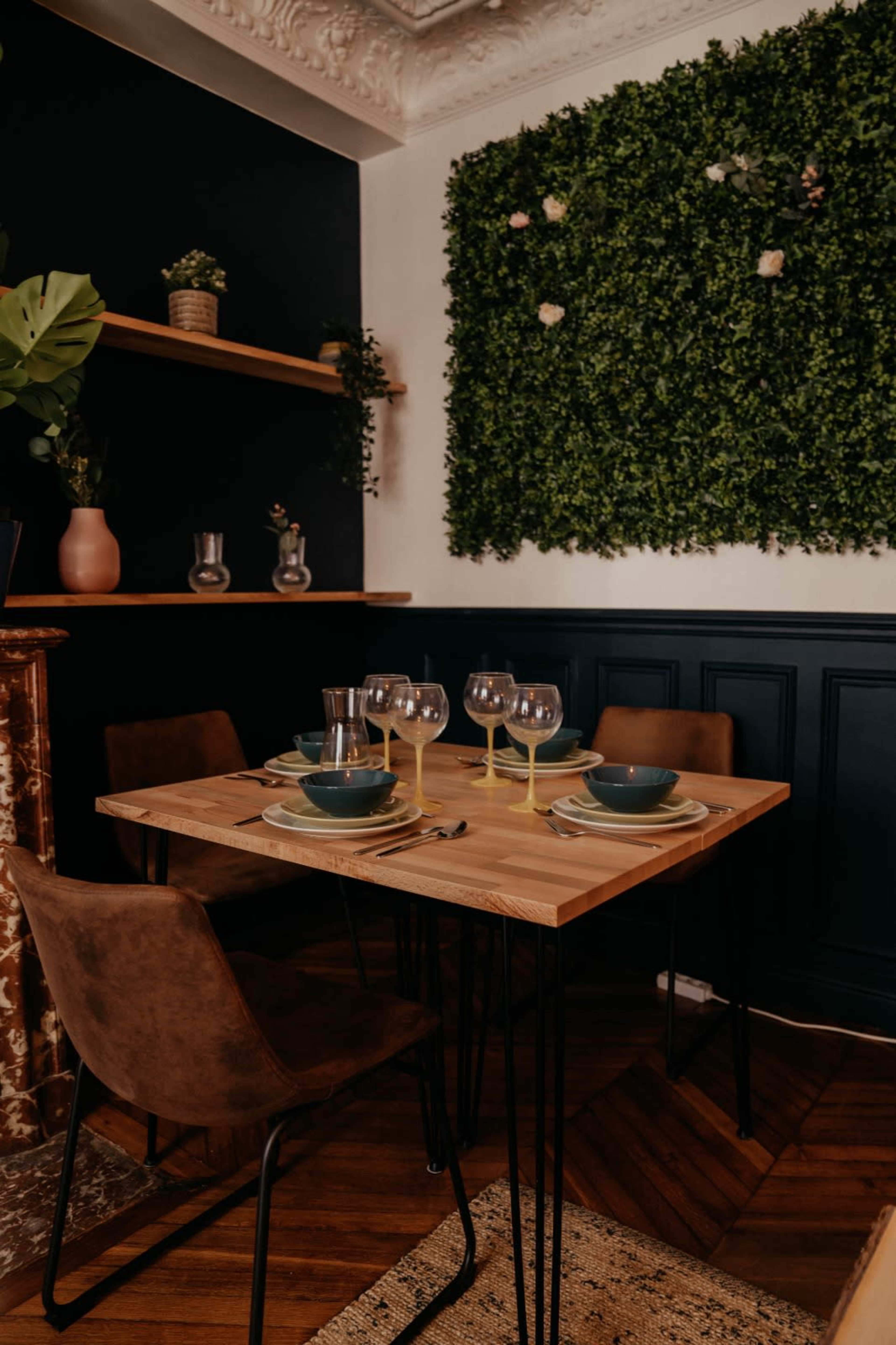 A dining table is set with plates and glasses in a room featuring a dark green plant wall and wooden accents.