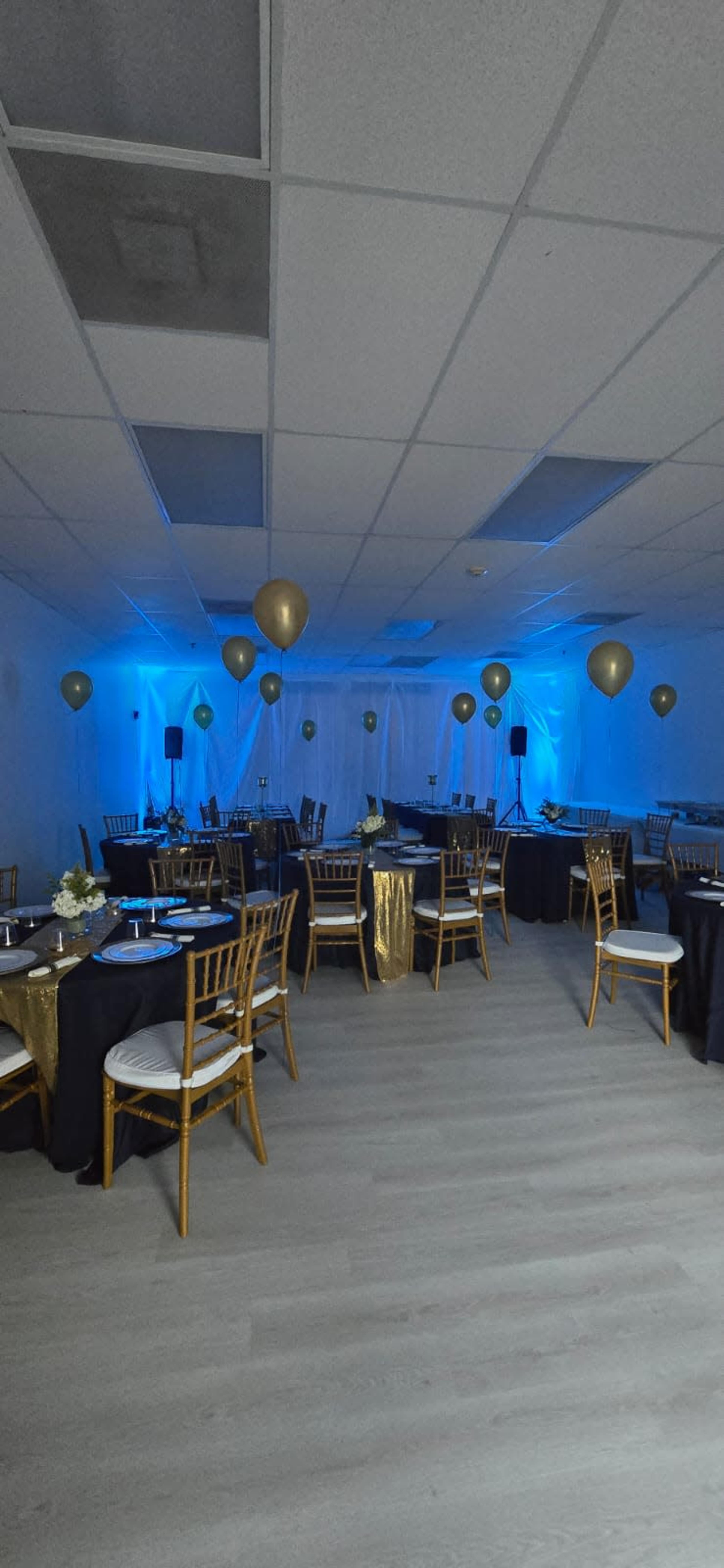 A banquet room is arranged with black and gold tablecloths, chairs, and balloons, featuring blue lighting and a minimalist decor.