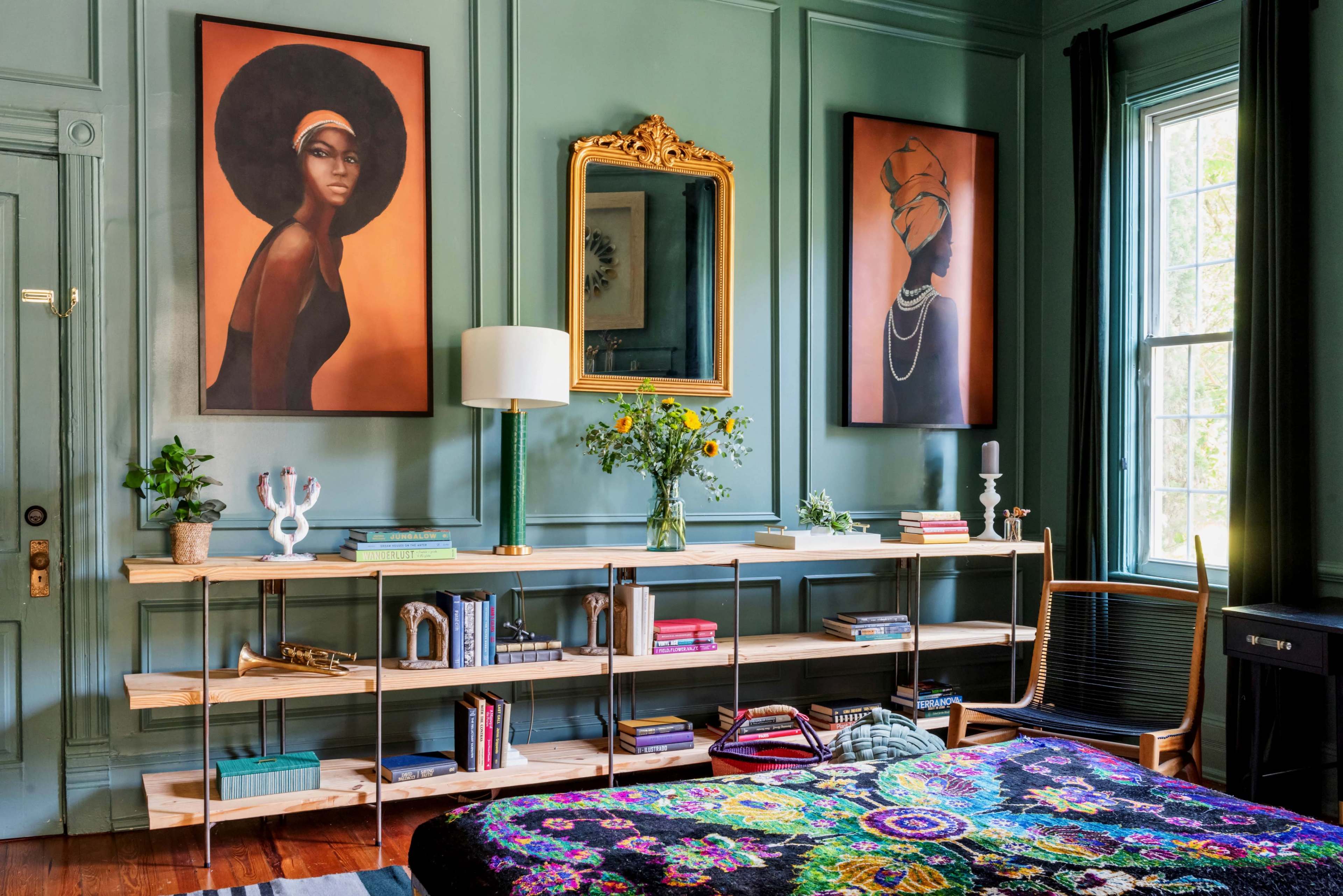 The image shows a stylish room with green walls, featuring two large framed portraits, a wooden shelf with books and decorative items, a bed with a colorful blanket, and a window letting in natural light.