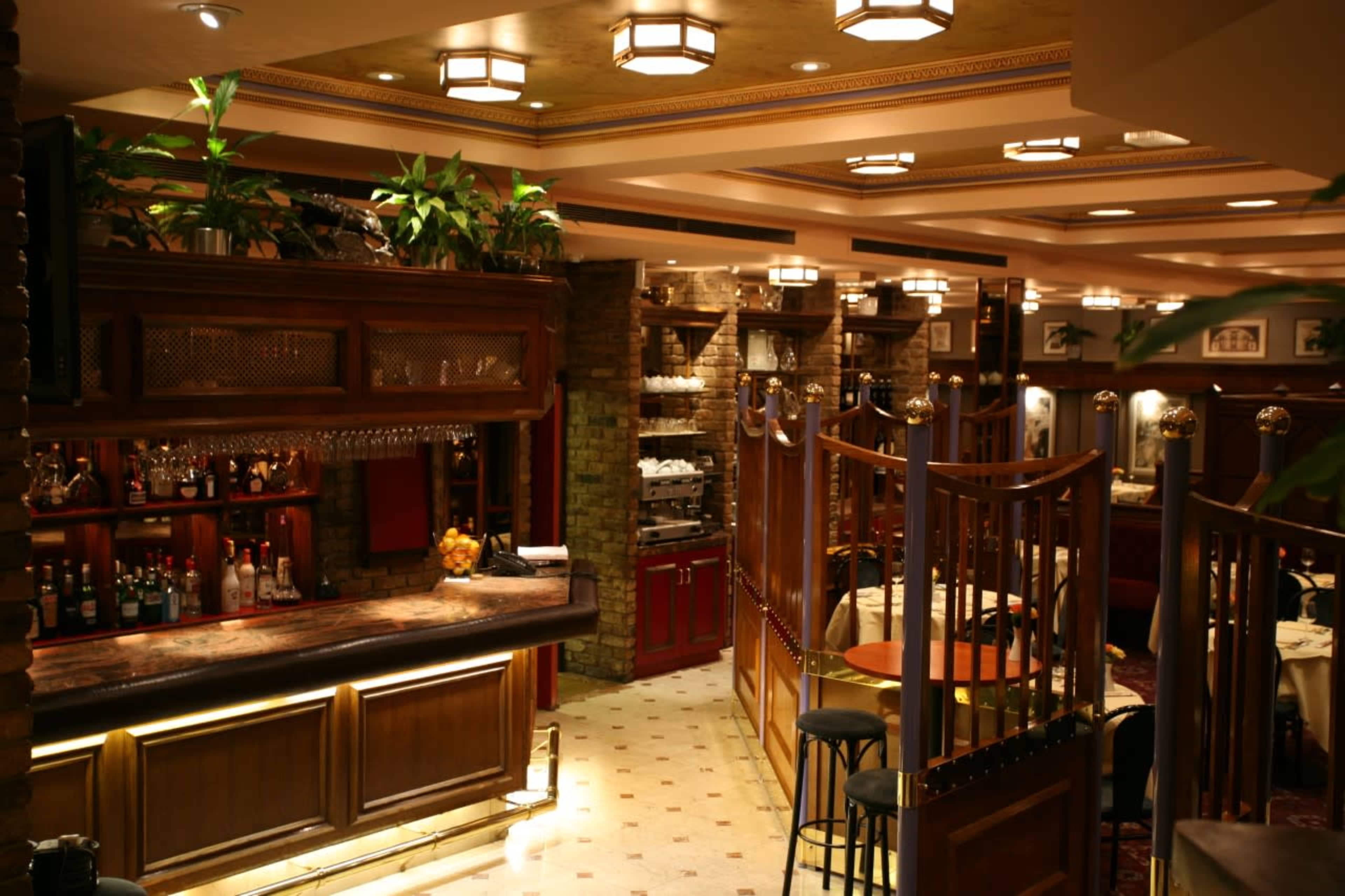 An elegant restaurant interior featuring a wooden bar, various plants, and separate dining areas with round tables and chairs.
