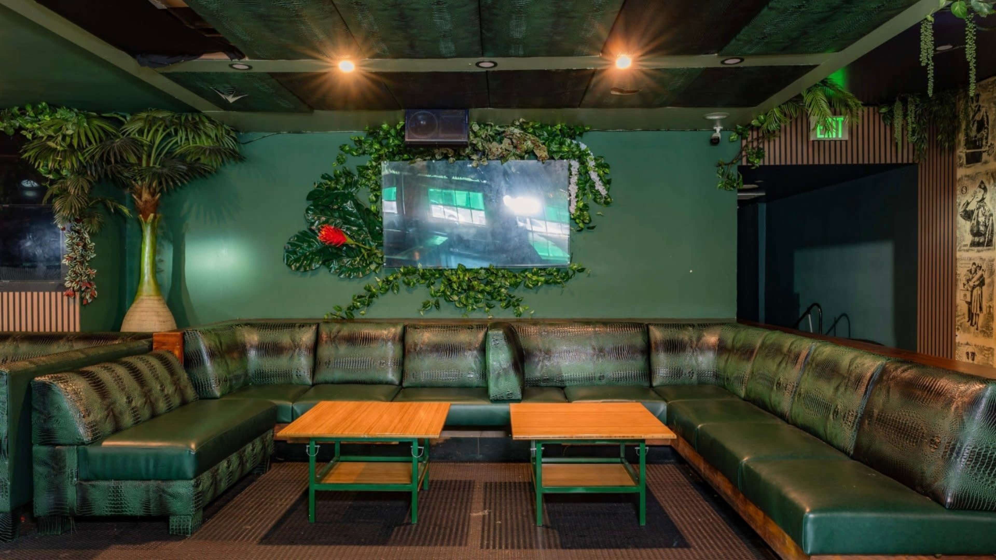 The image shows a lounge area featuring a green leather sectional sofa, two wooden coffee tables, and a large wall-mounted TV surrounded by decorative plants.