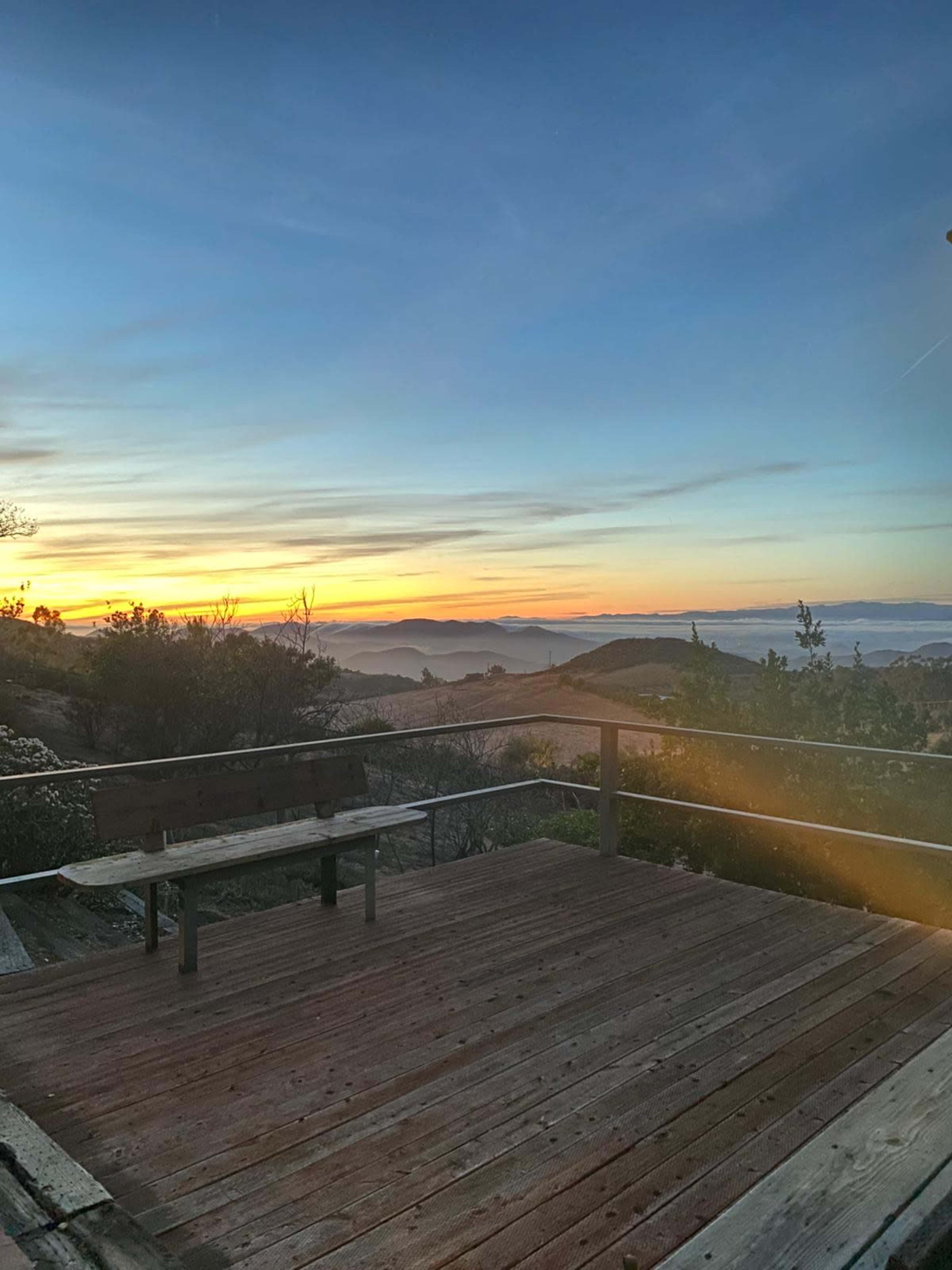 A wooden deck overlooks a mountainous landscape at sunset, with vibrant colors in the sky and a visible horizon.
