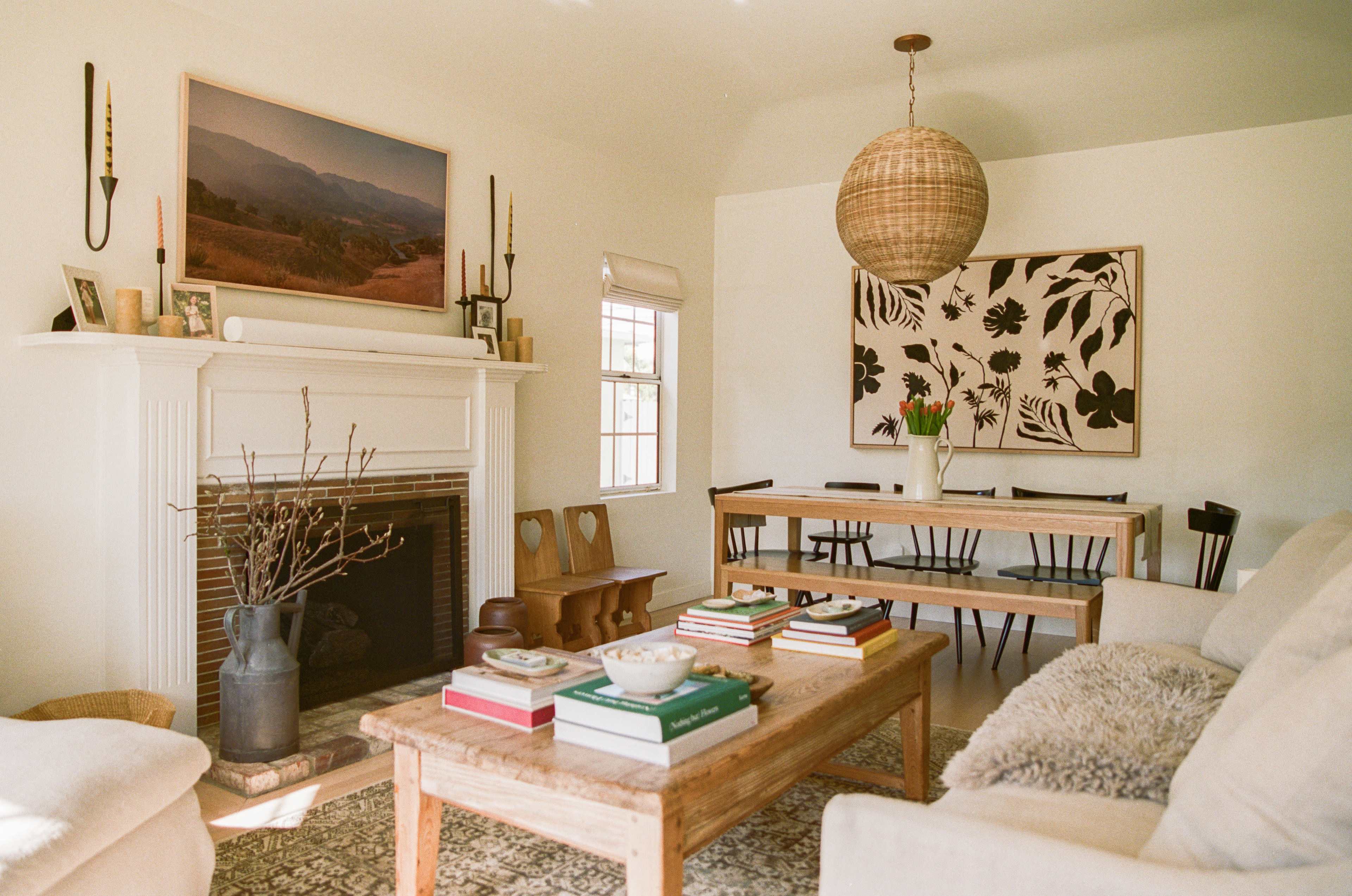A bright living room featuring a fireplace, a wooden coffee table with stacked books, a dining table, and a large wall art piece.