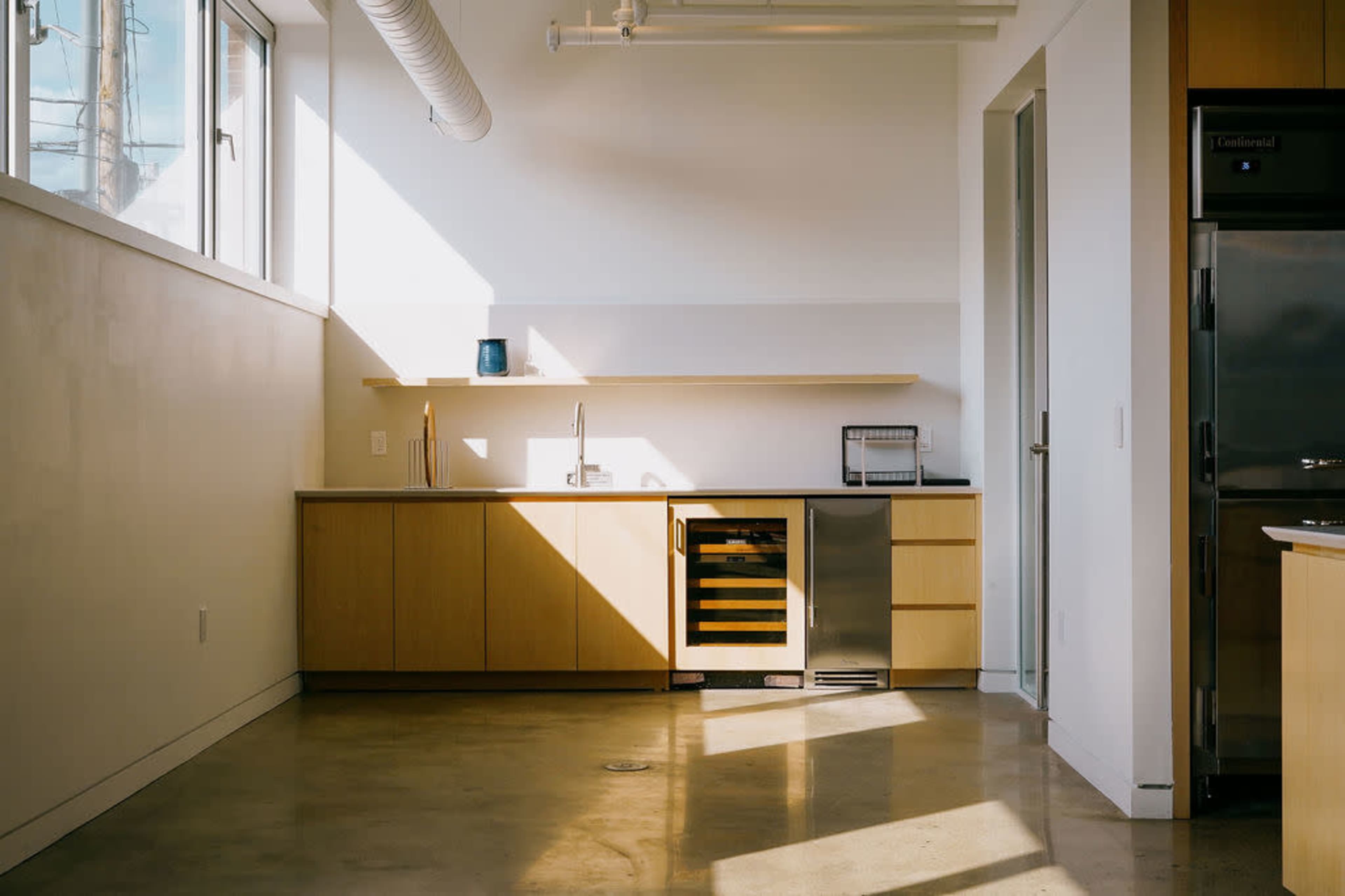 A modern kitchen features wooden cabinetry, a wine cooler, a stainless steel refrigerator, and large windows allowing natural light to enter.