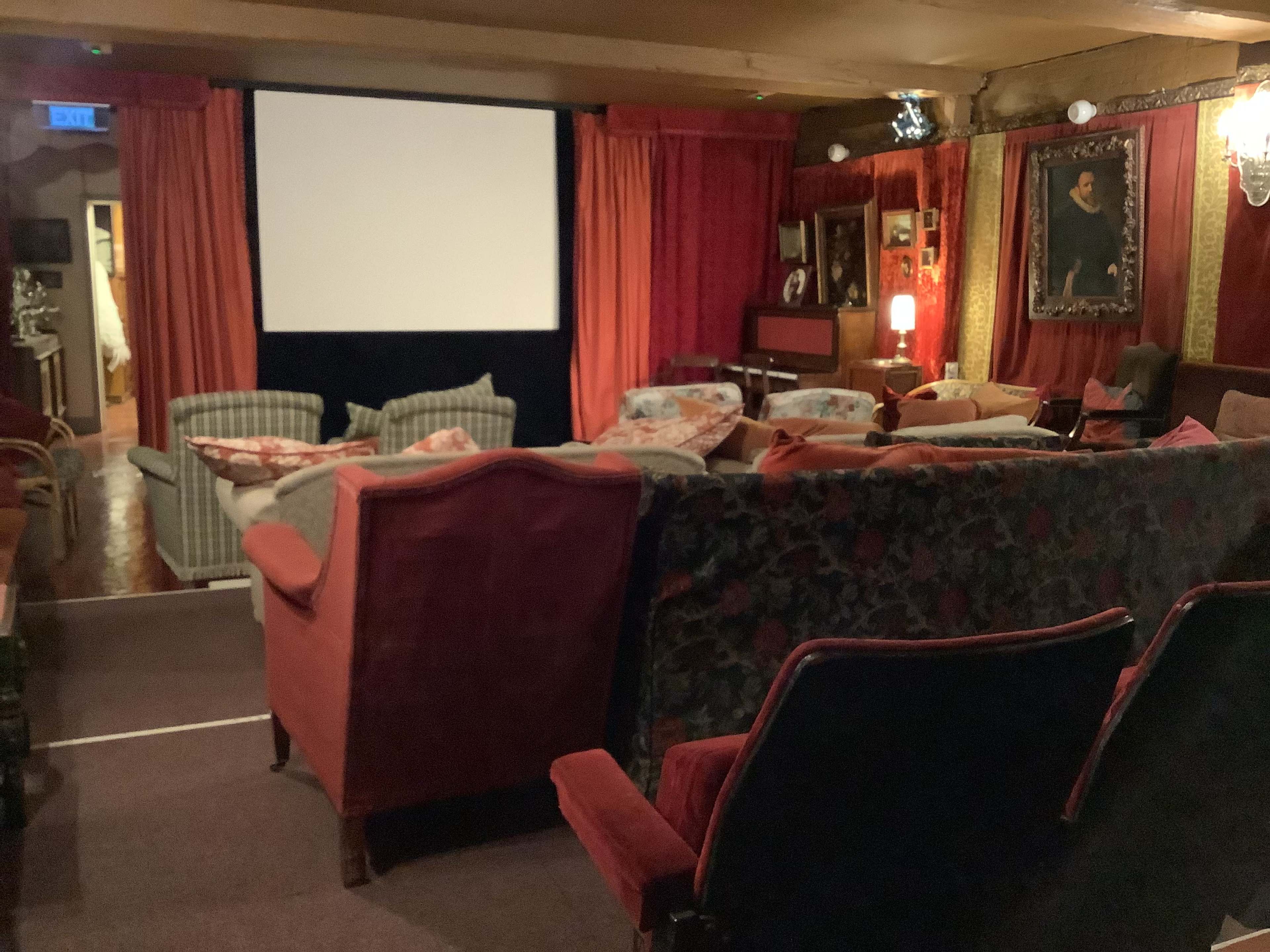 A cozy, vintage-style cinema room with plush seating facing a blank projector screen, surrounded by rich curtains and framed artwork on the walls.