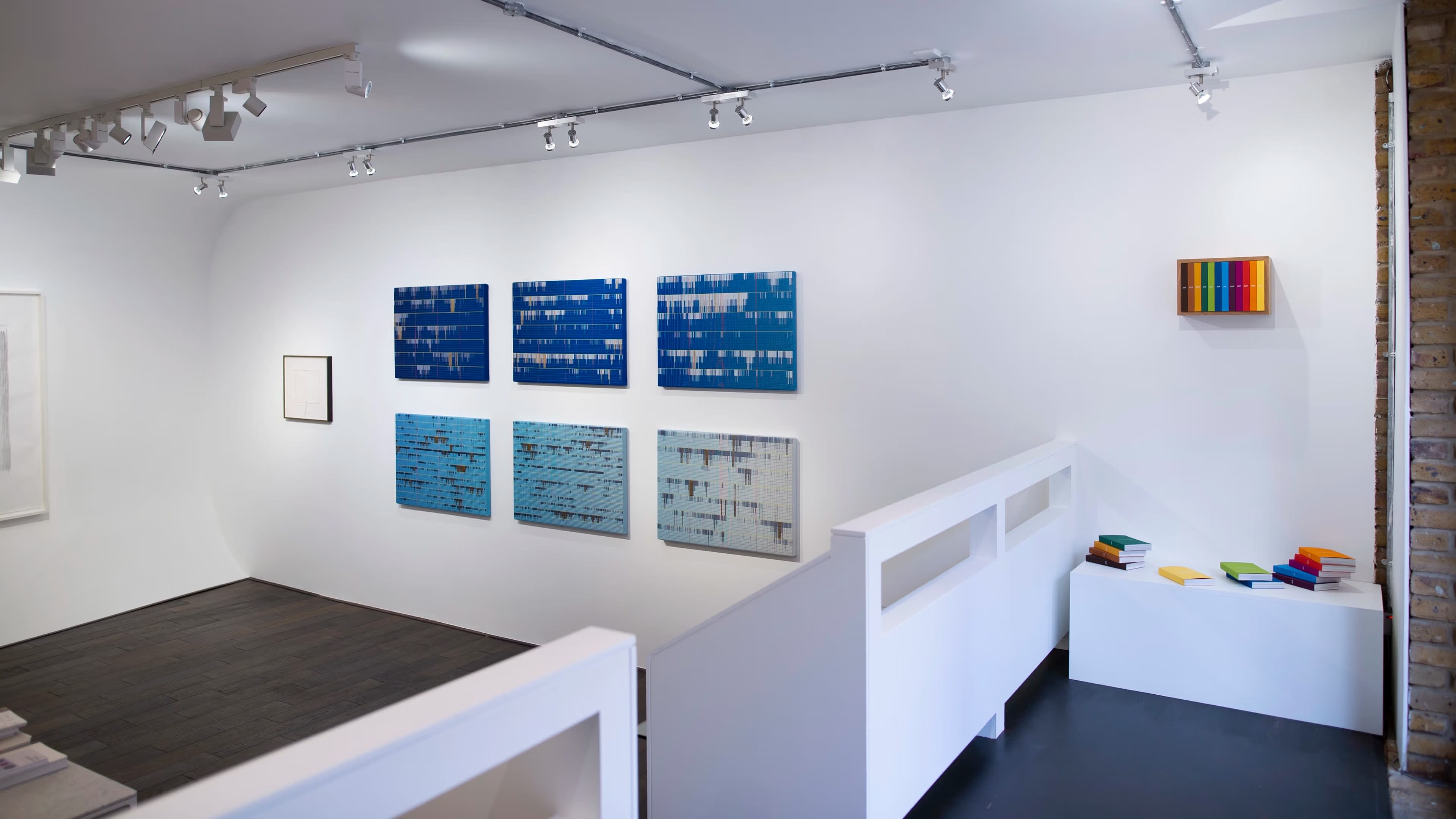 The image shows a minimalistic art gallery interior with several abstract blue artworks displayed on the walls, a small stack of colorful books or objects in one corner, and a bright white railing separating different areas of the space.