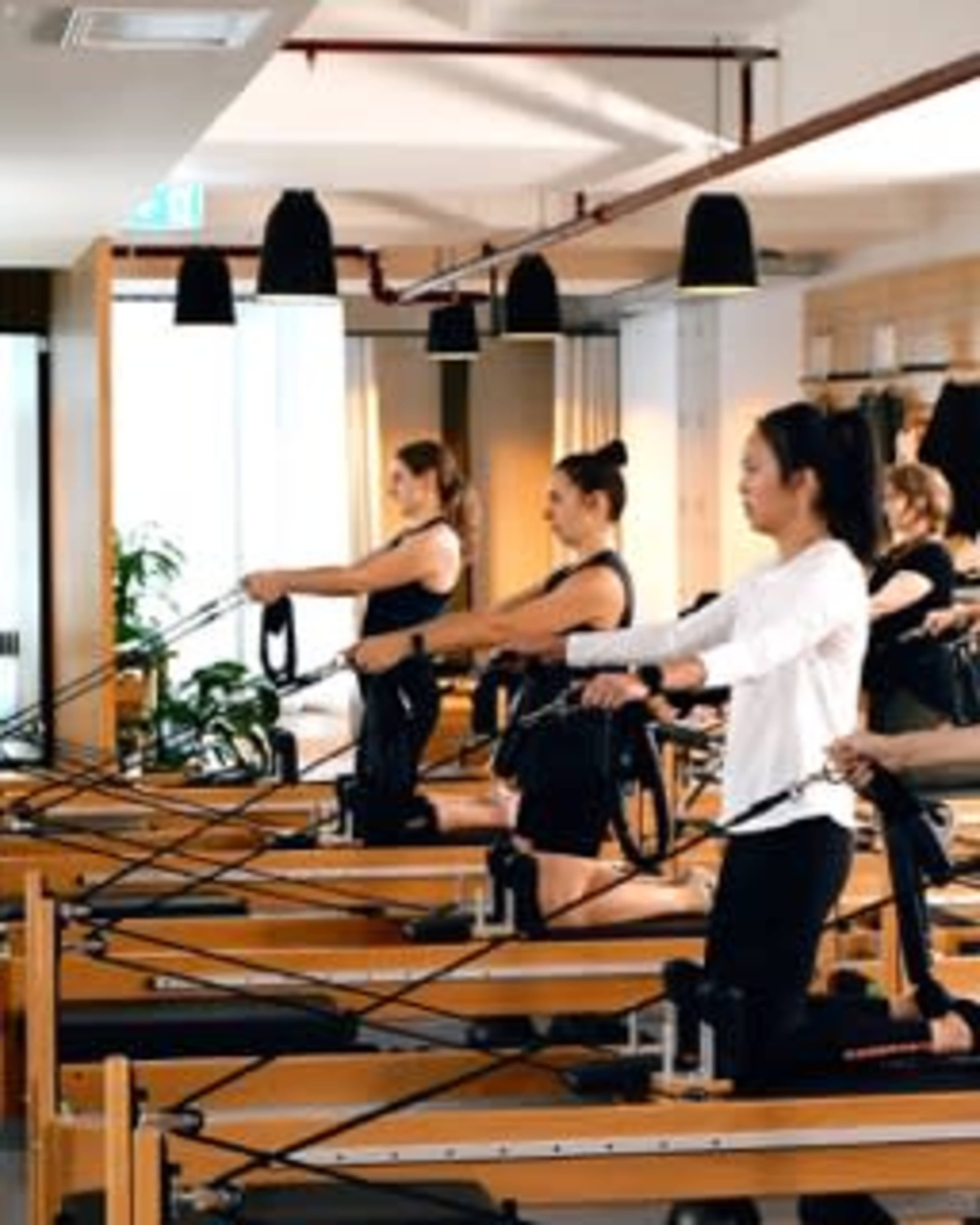 A group of individuals is performing exercises on reformer Pilates machines in a bright studio.