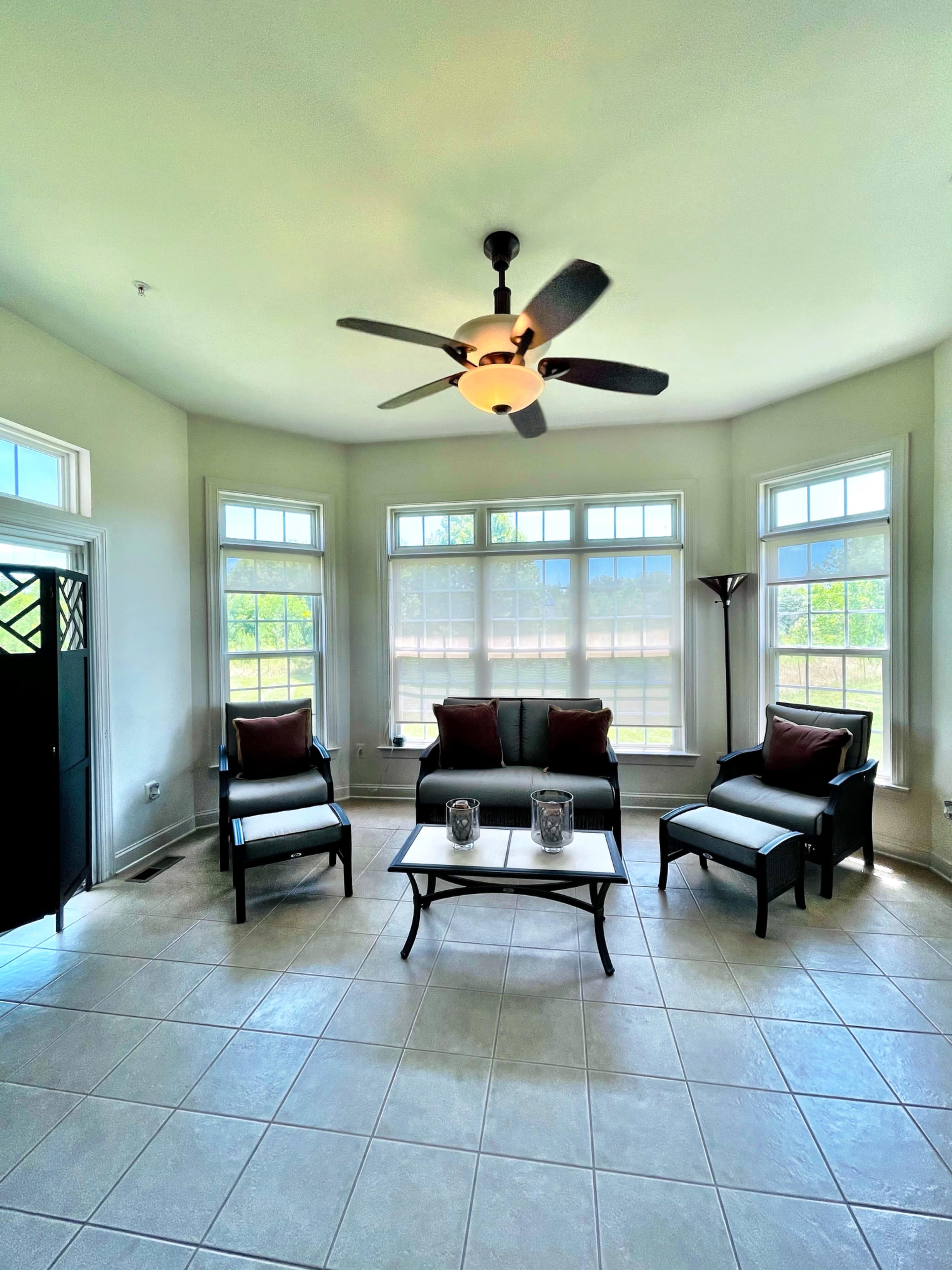 A sunlit room with large windows, a ceiling fan, a sofa, and two chairs arranged around a coffee table.