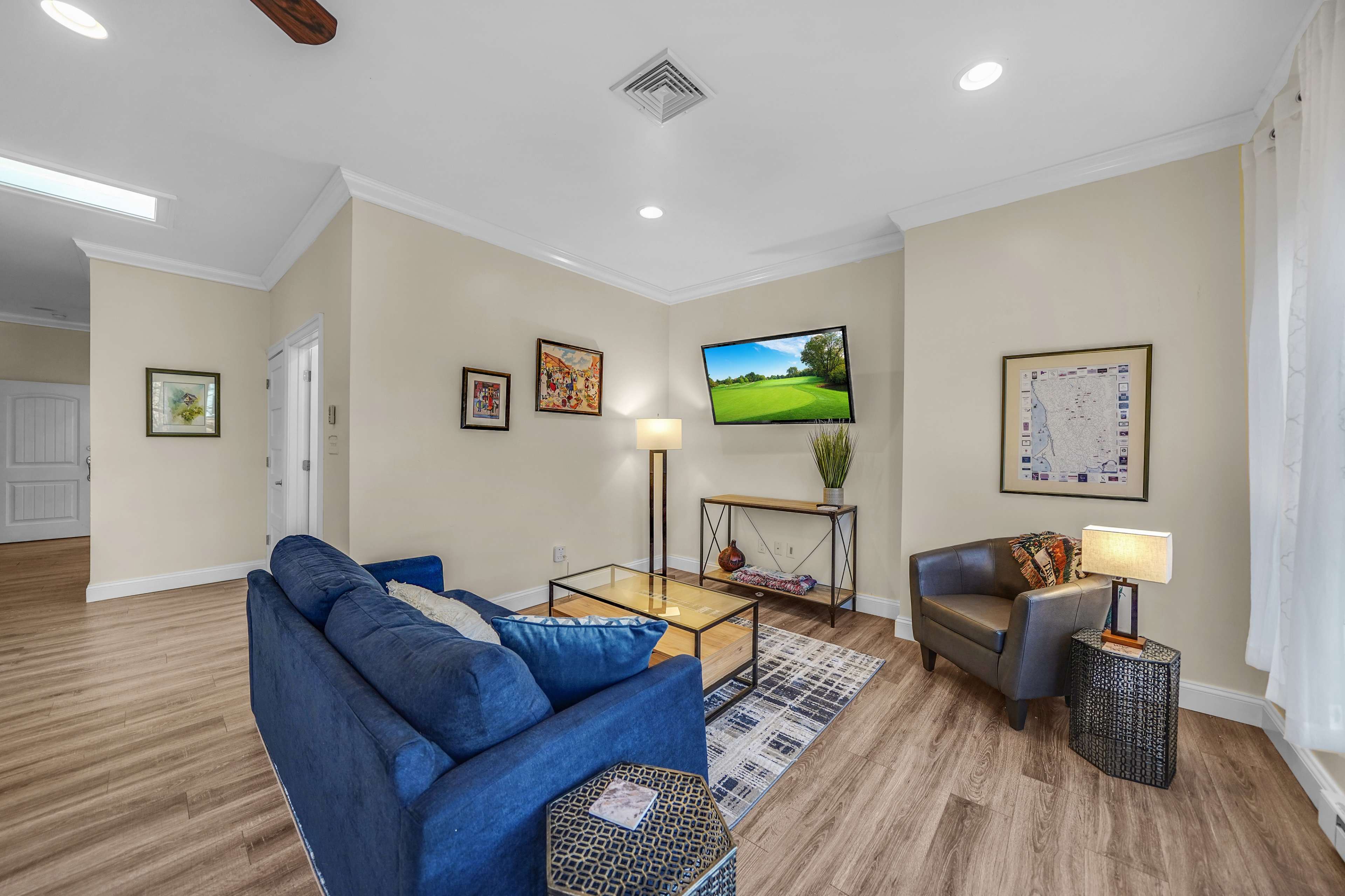 The living room features a blue sofa, a glass coffee table, and a television mounted on the wall, with framed artwork and a decorative lamp.