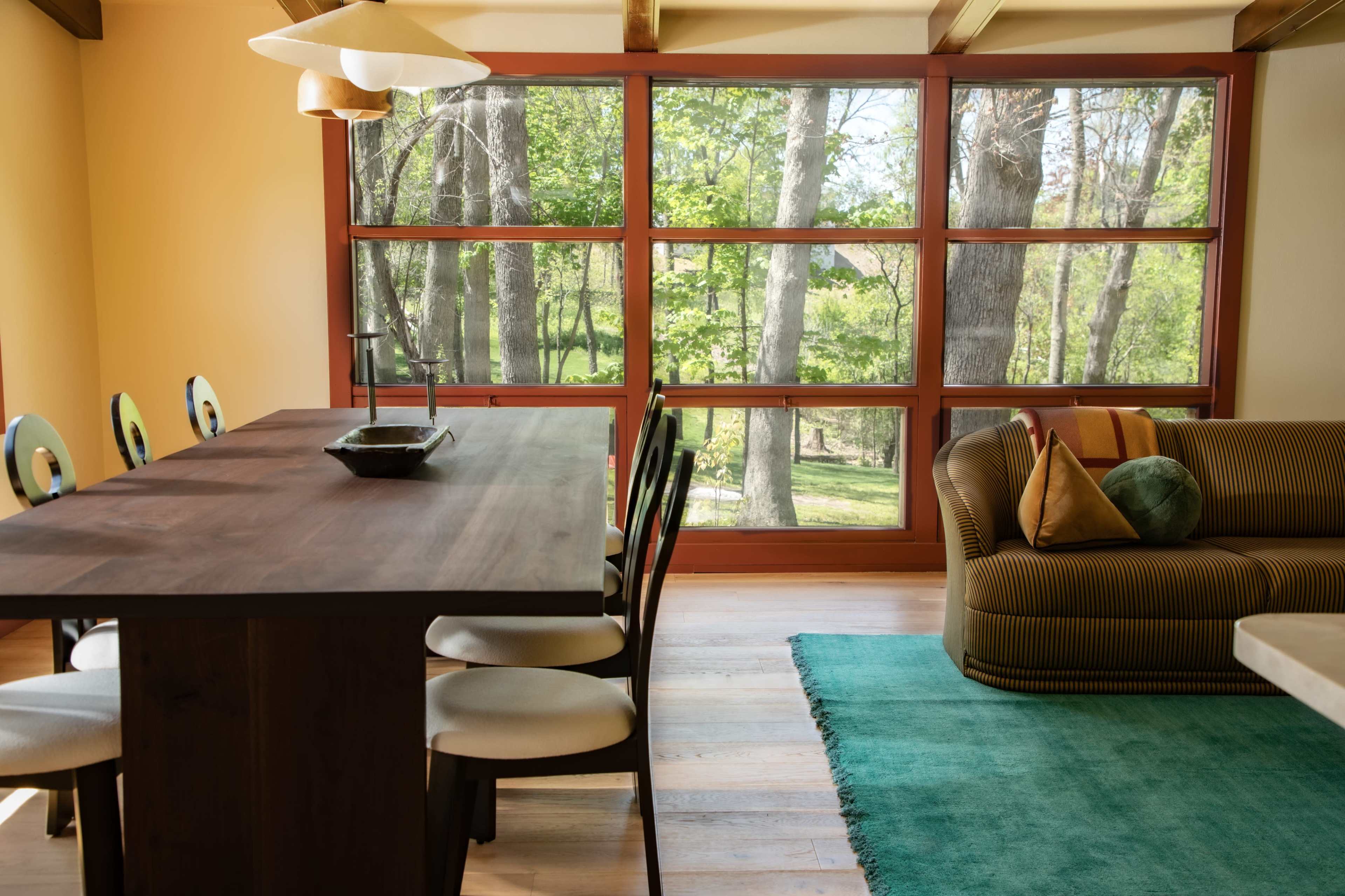 A dining area features a wooden table with black chairs, a green area rug, and a sofa, all alongside large windows offering a view of trees outside.