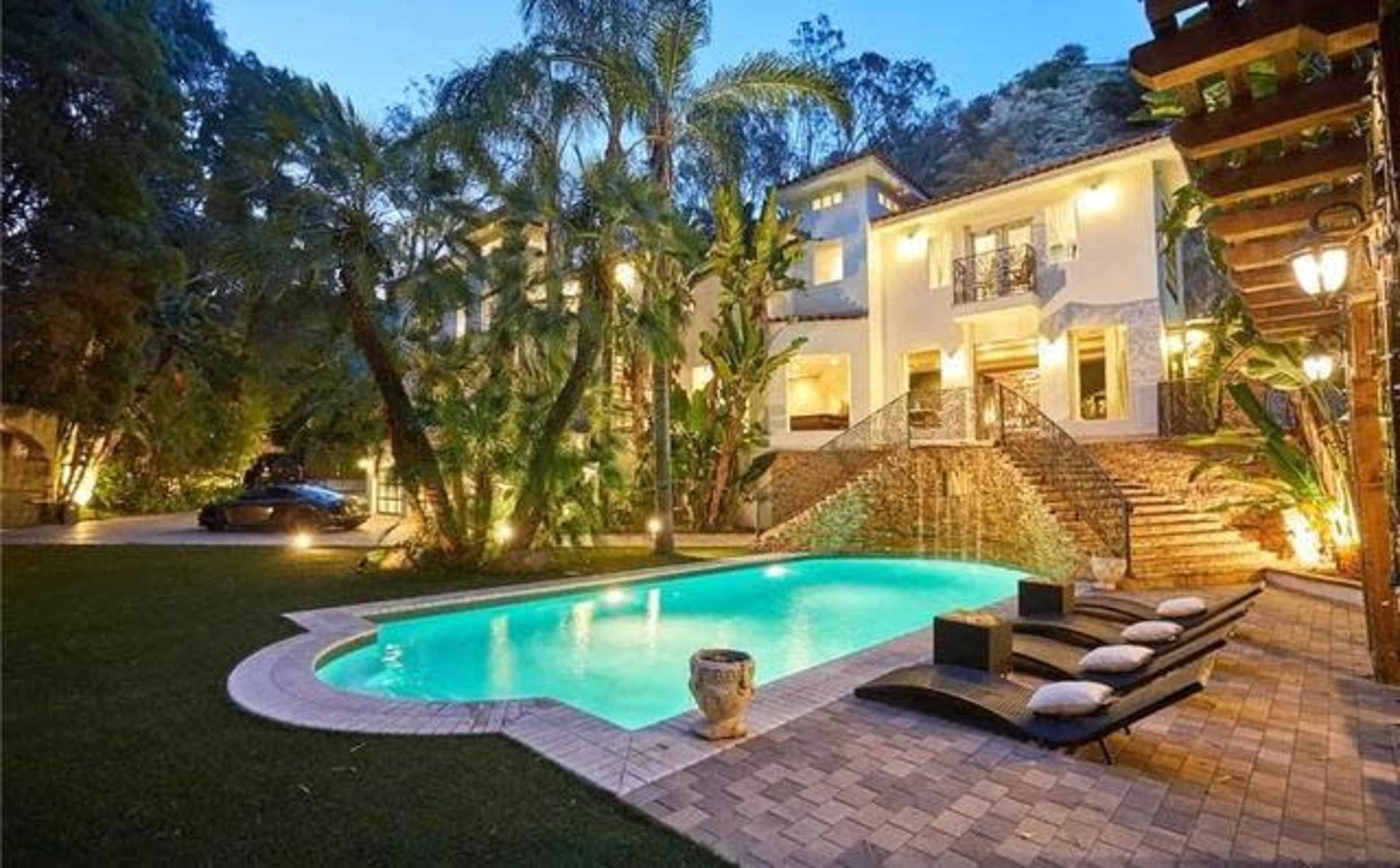 The image shows a well-lit modern mansion with a swimming pool surrounded by palm trees and lounge chairs in the backyard.