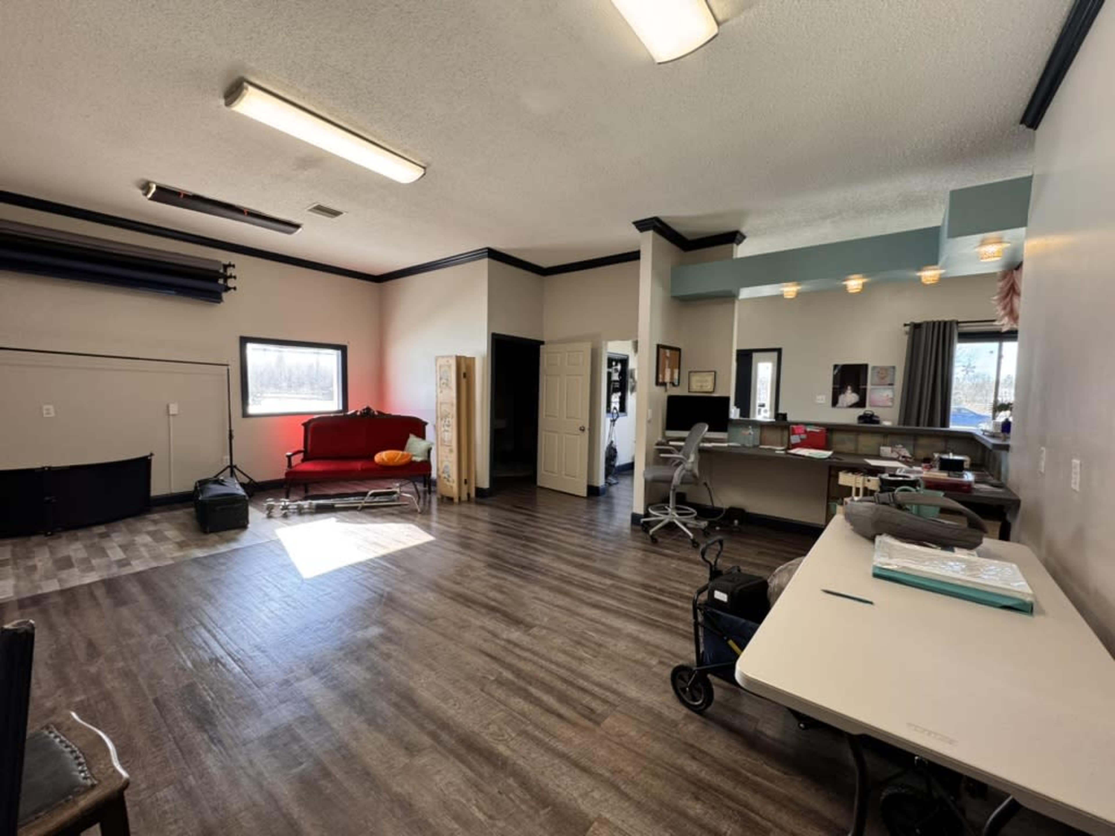 The image shows a spacious room with a red couch, wooden flooring, a desk area, and large windows allowing natural light to enter.