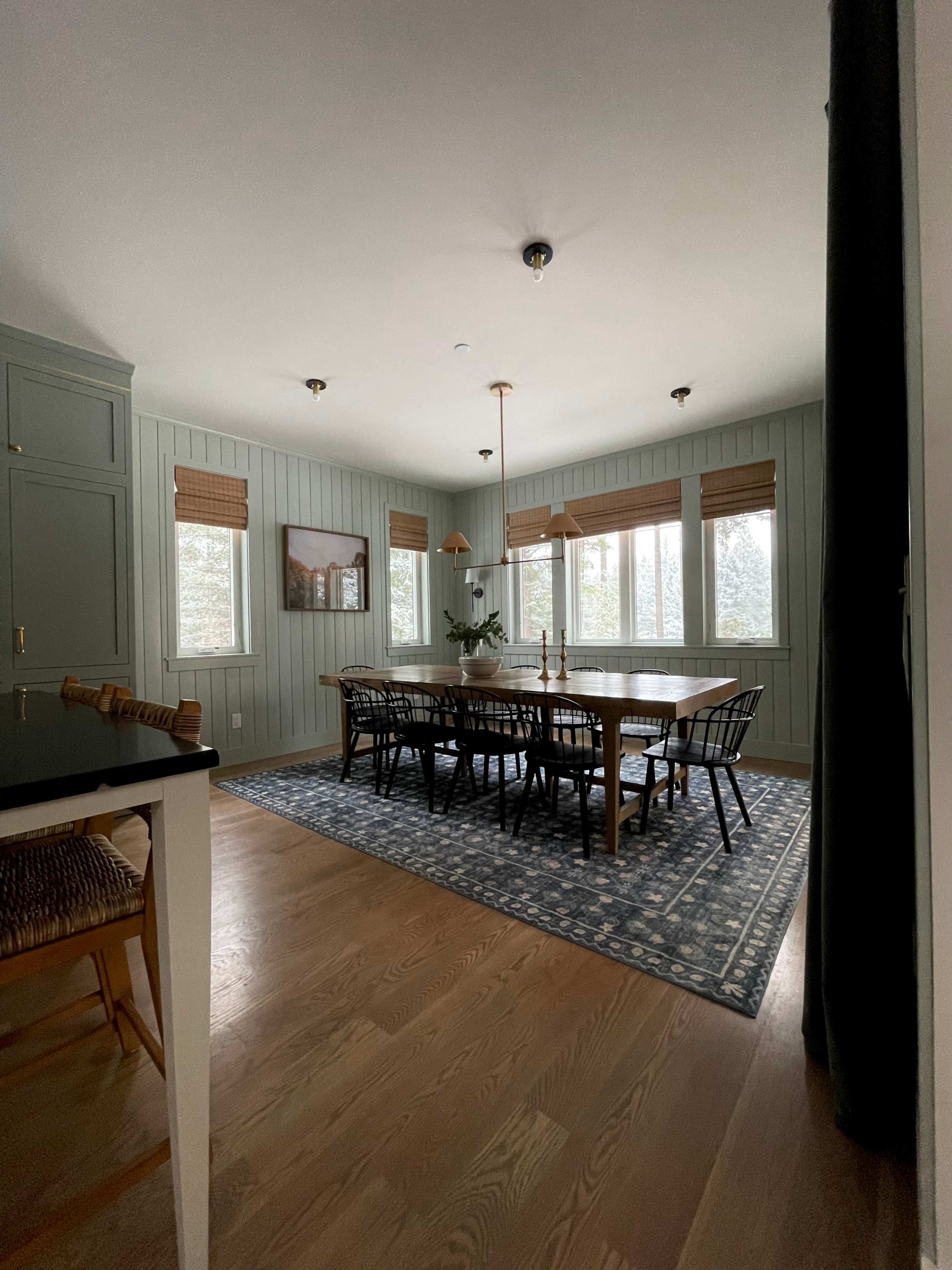 A spacious dining room features a large table surrounded by black chairs, with a patterned rug on the hardwood floor and natural light filtering through large windows.