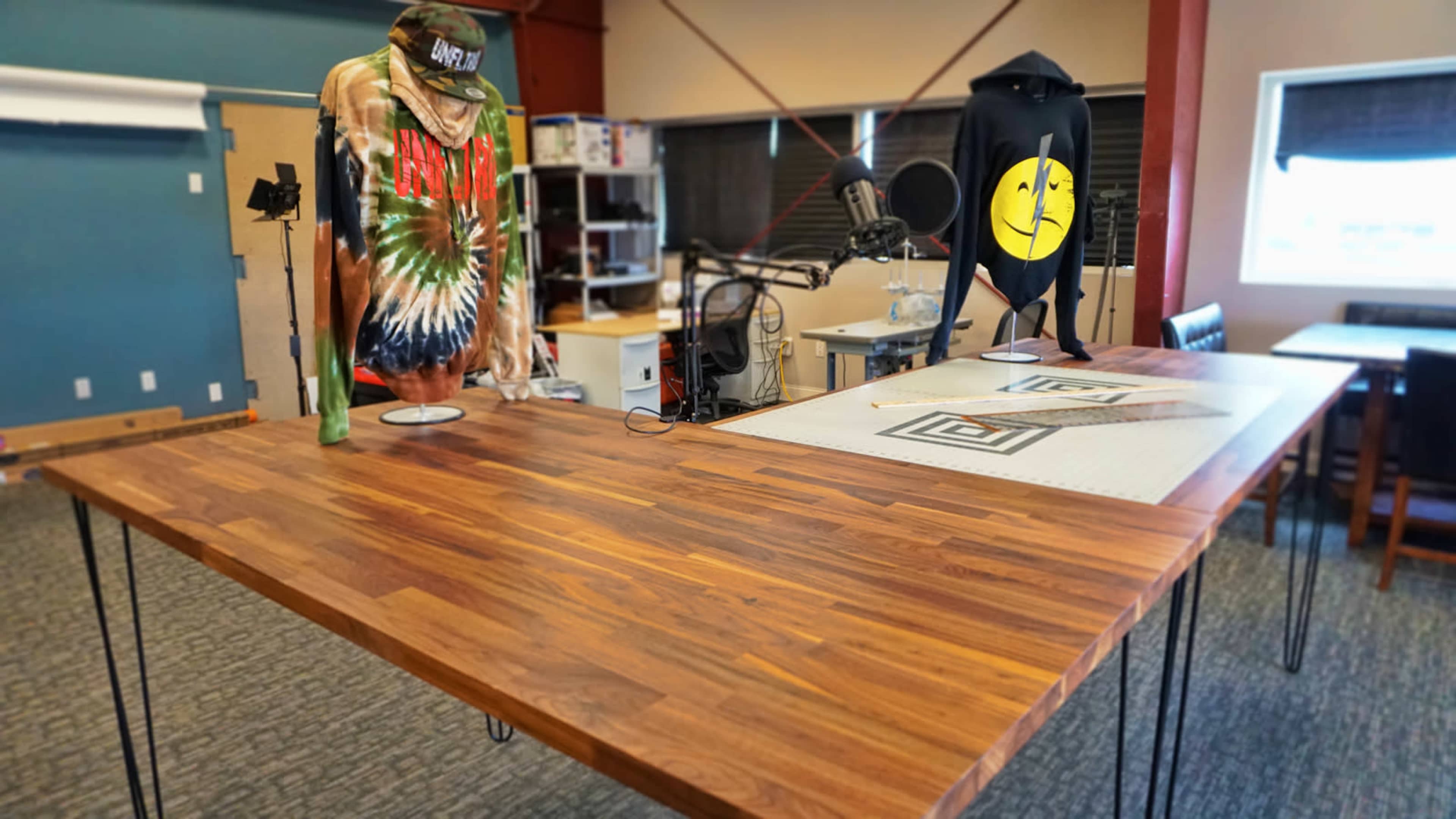 The image shows a spacious room with a wooden table featuring a tie-dye shirt and a hoodie displayed on stands, alongside a microphone setup.