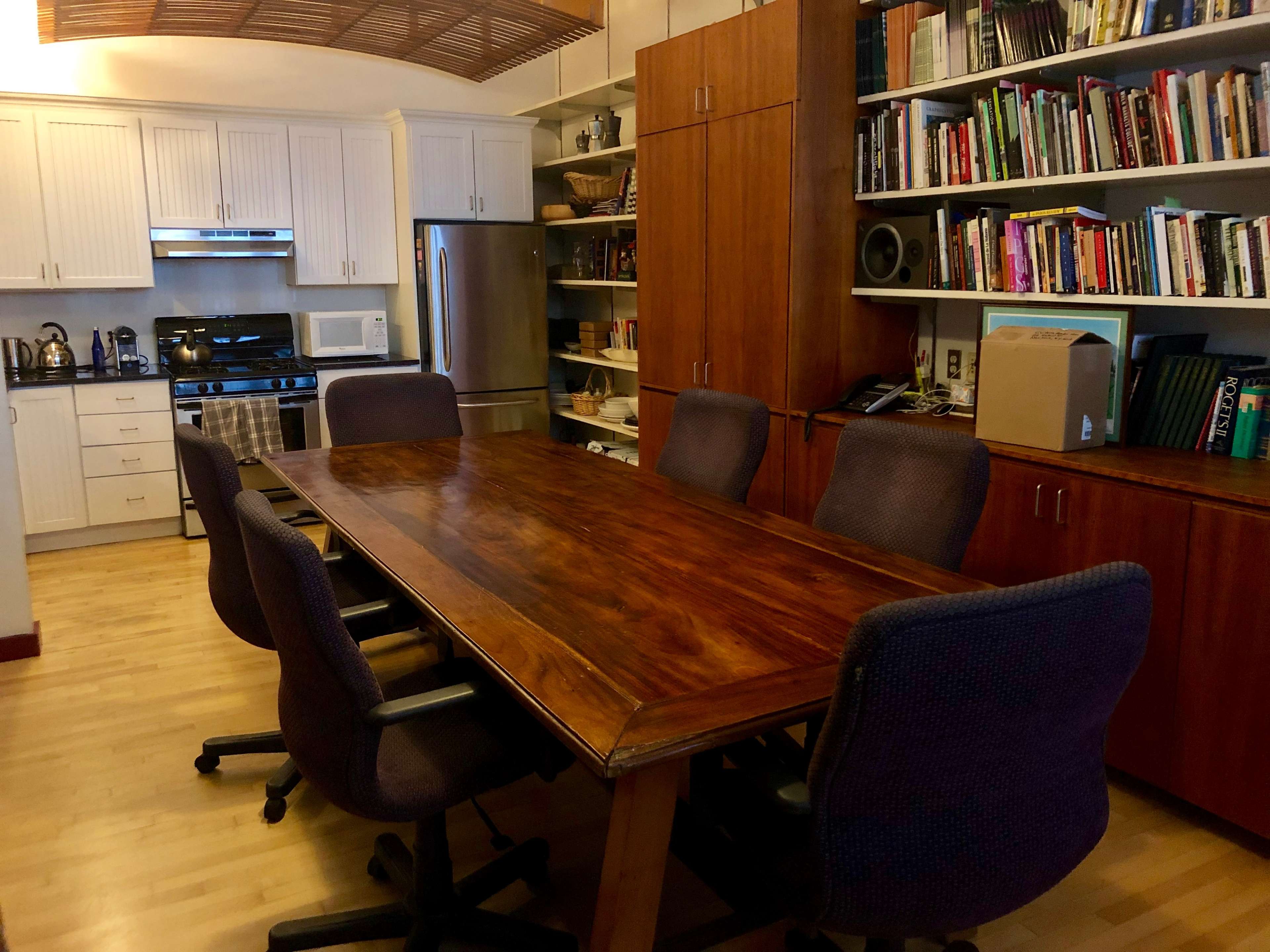 A large wooden conference table surrounded by five ergonomic chairs, situated in a kitchen-like space with a modern kitchen area and bookshelves.