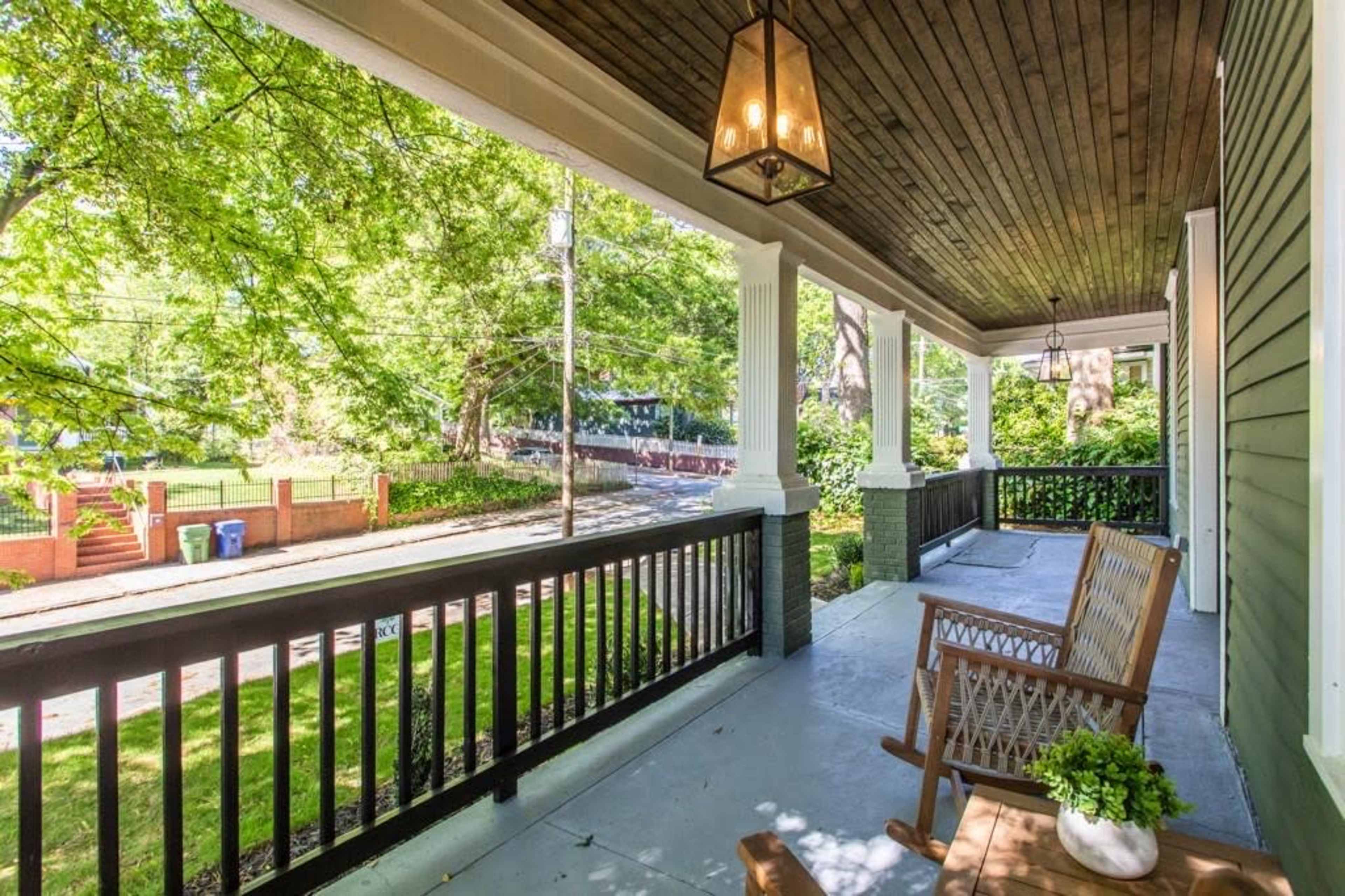 A porch with a wooden bench and a small table, surrounded by greenery and a pathway leading to a street.