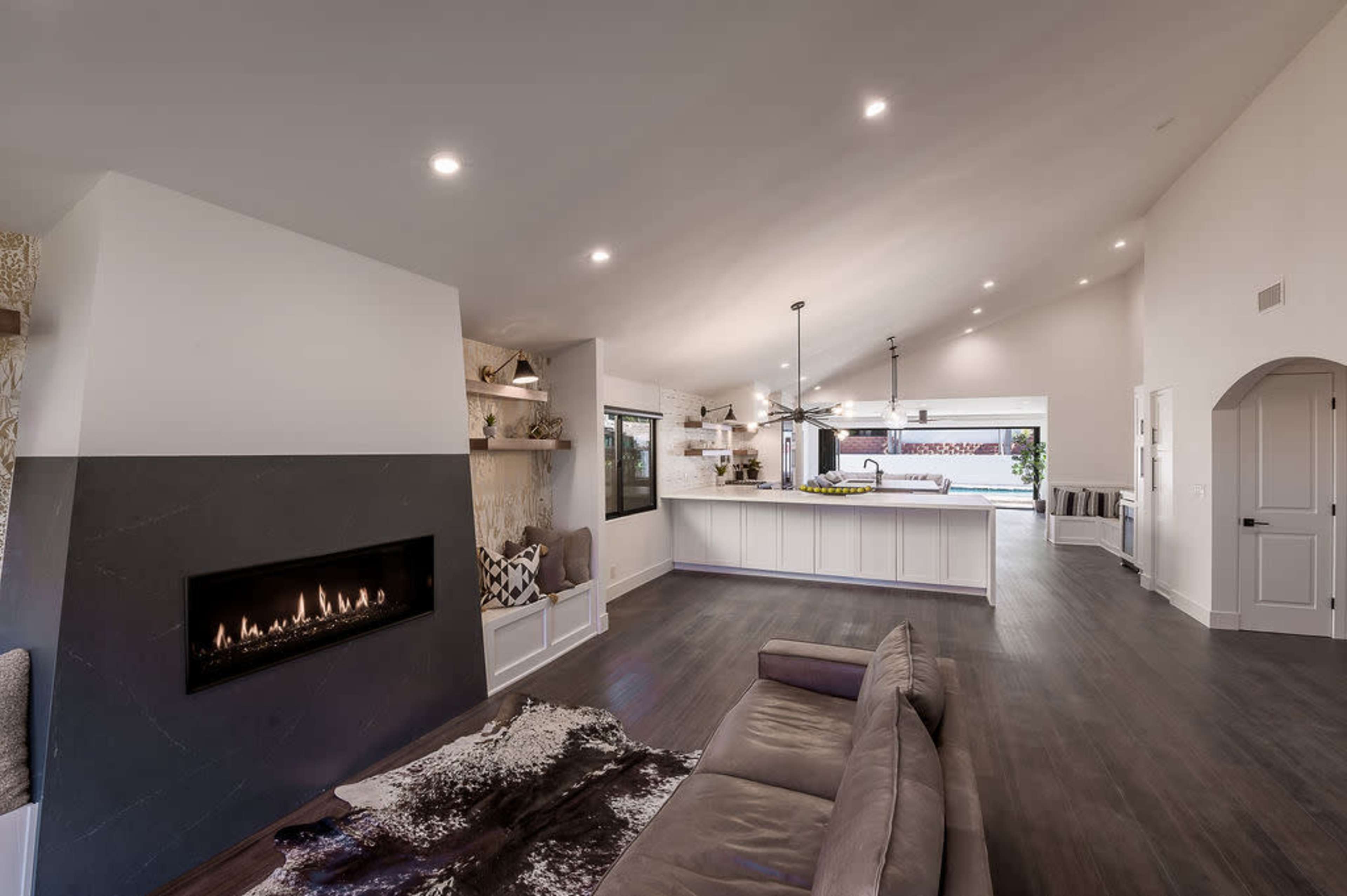 The image shows a modern living room with a fireplace, a large open kitchen, and an area with neutral furniture and hardwood flooring.