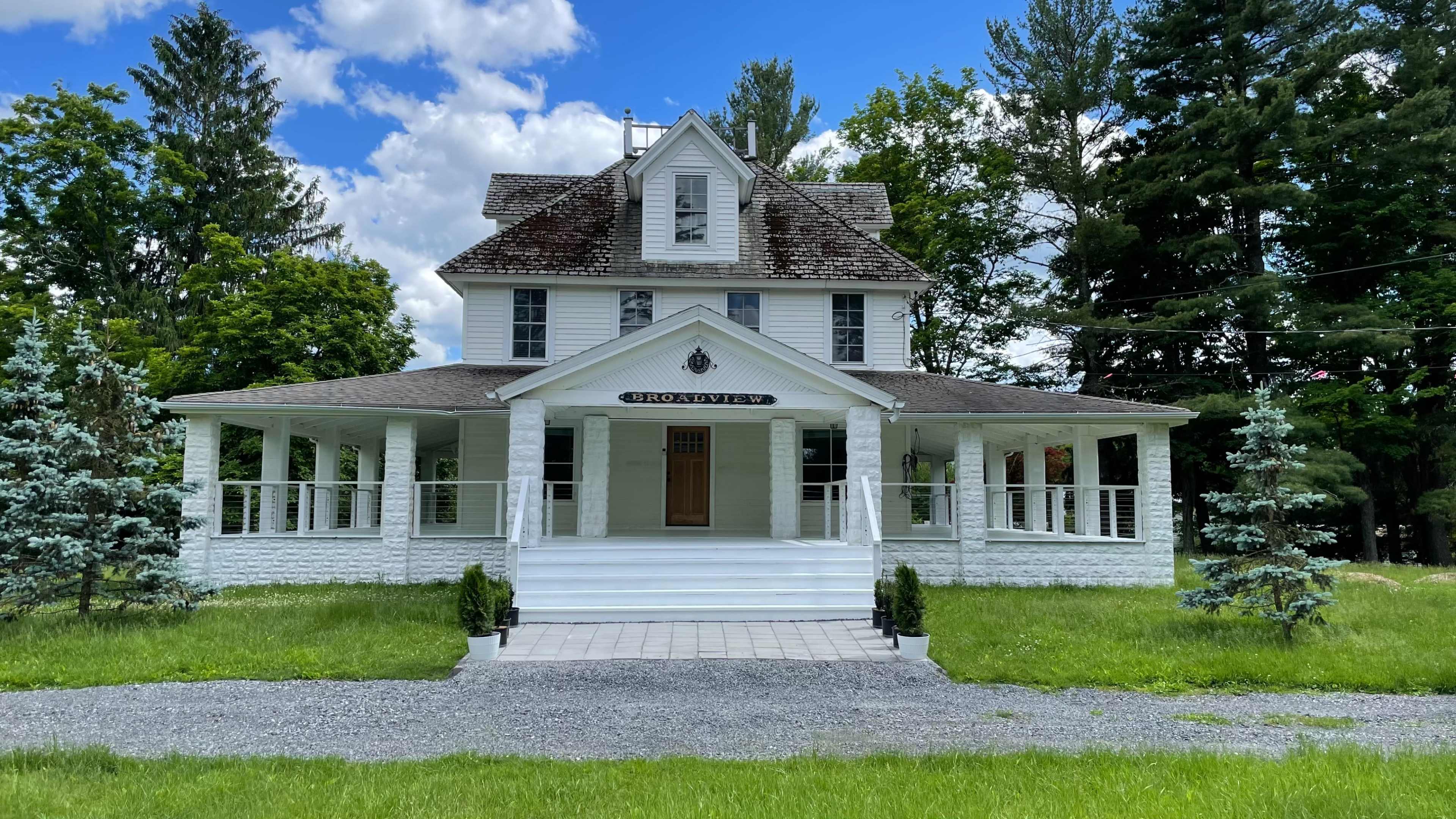 A large, white house with a wraparound porch, set on a grassy area with a gravel driveway under a blue sky dotted with clouds.