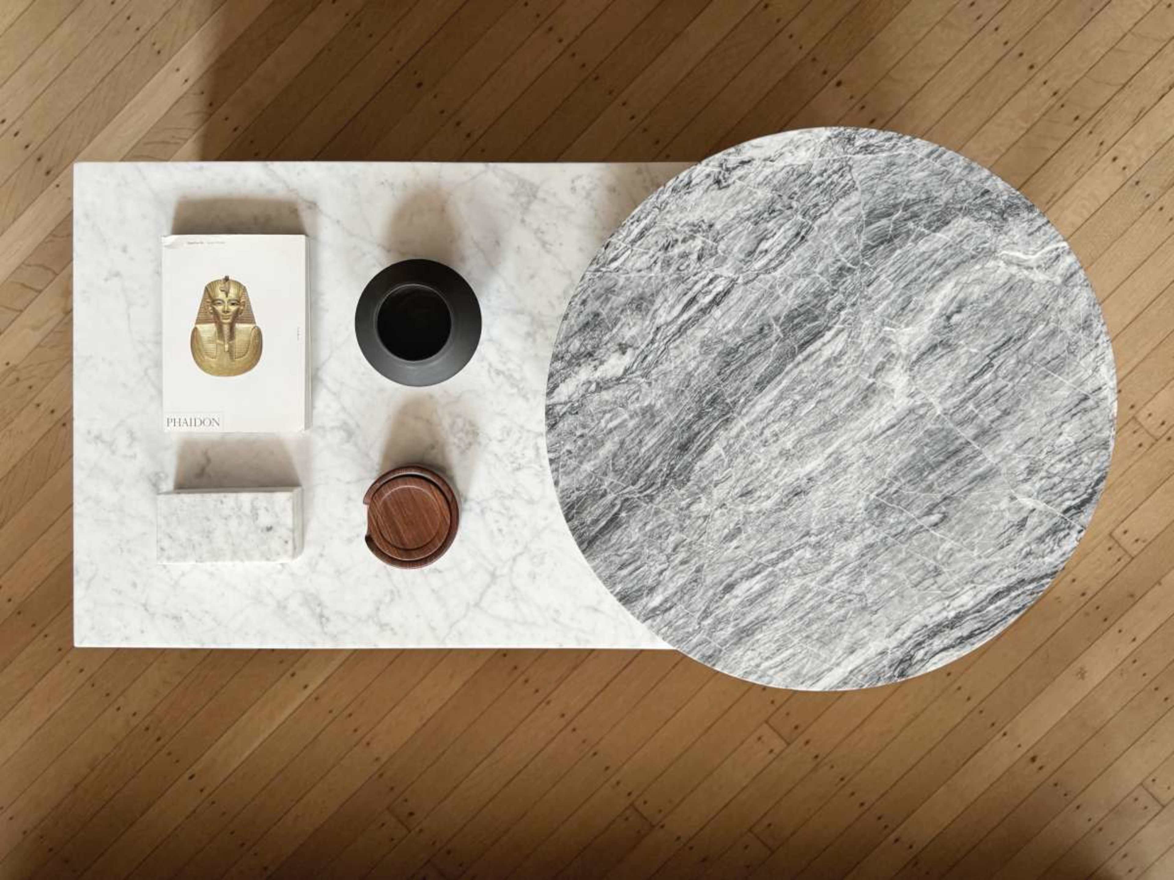 The image shows a marble table with a circular top, featuring a book, a small black vase, and a wooden container.