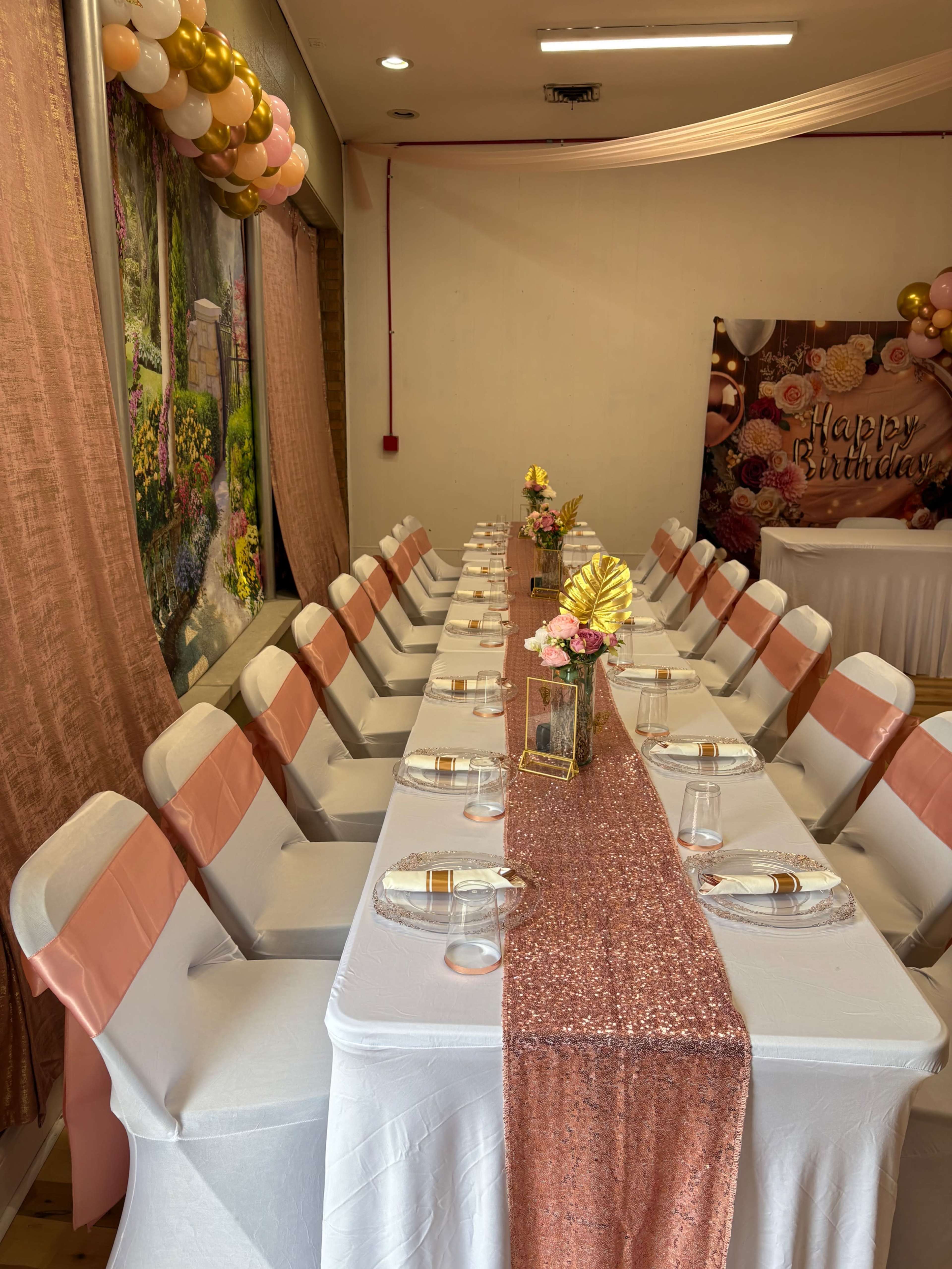 The image shows a long banquet table elegantly set with white tablecloths, pink accents, and decorative floral centerpieces in a decorated event space.