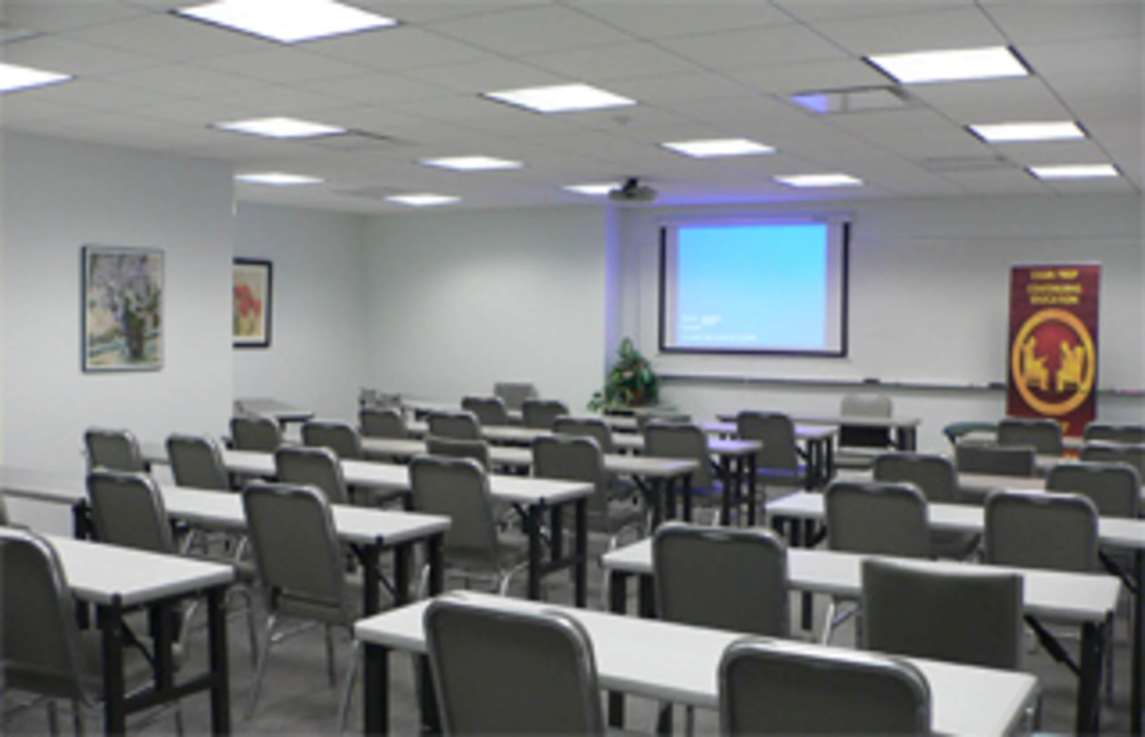 A classroom arranged with rows of tables and chairs, featuring a projector screen at the front and a banner on one side.