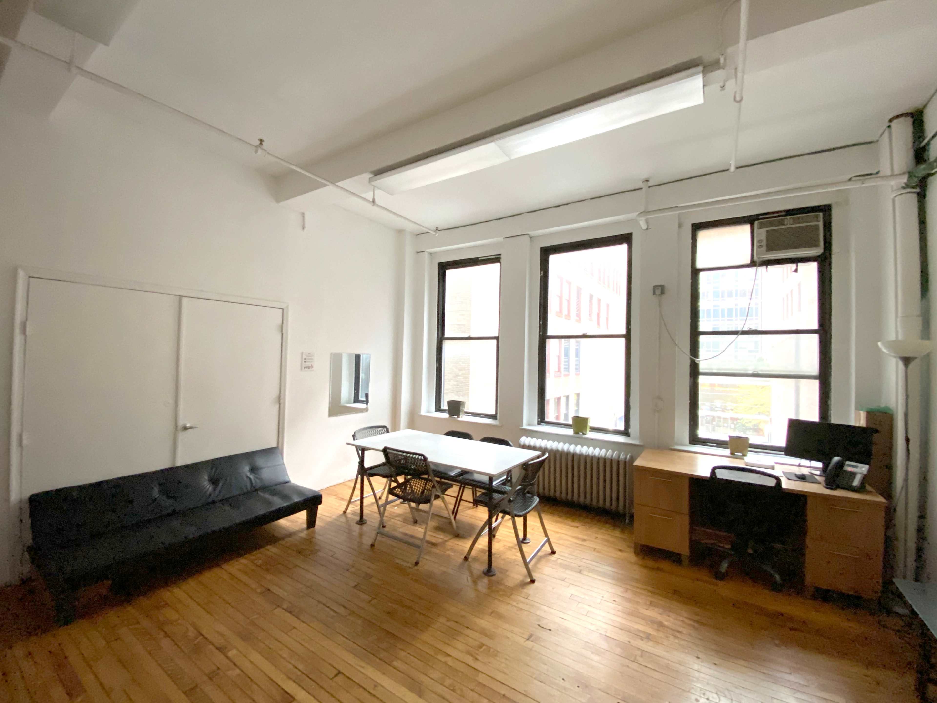 A spacious office with hardwood floors, two large windows, a black sofa, a table with folding chairs, and a desk with a computer.