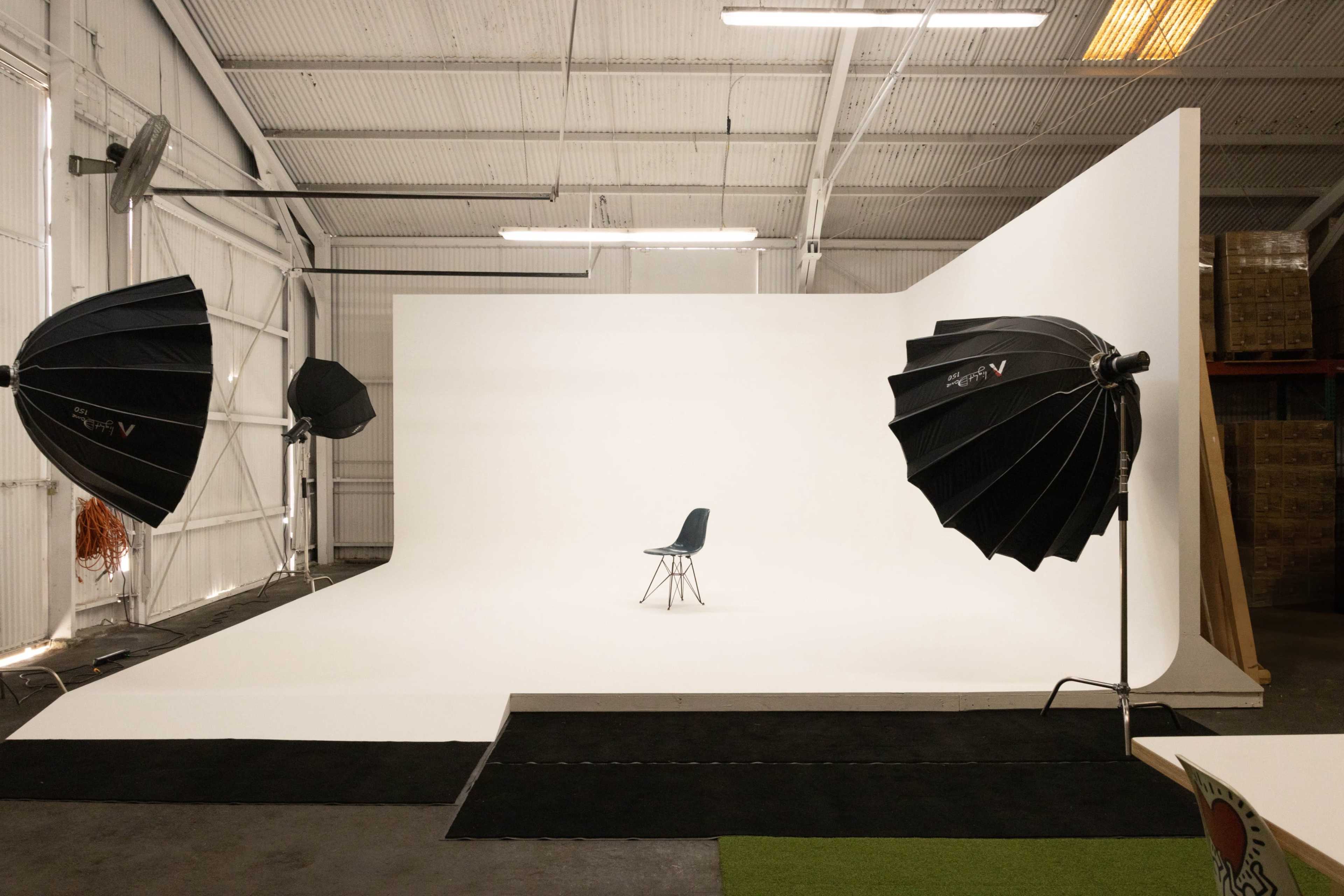 A simple photography studio features a white backdrop, a single chair at the center, and two large softbox lights on either side.
