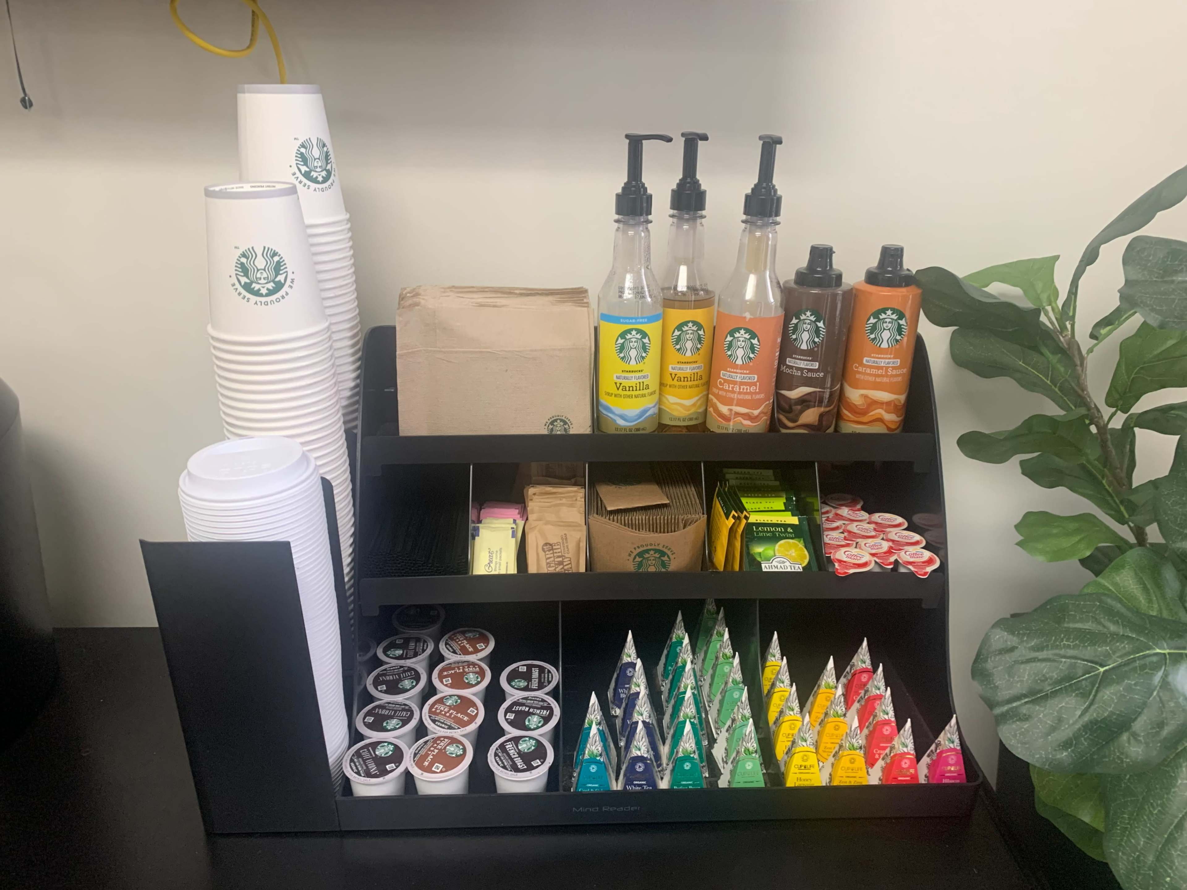 A beverage station featuring stacked Starbucks cups, various flavored syrups in dispensers, sugar packets, creamers, and tea bags organized on a black shelving unit.