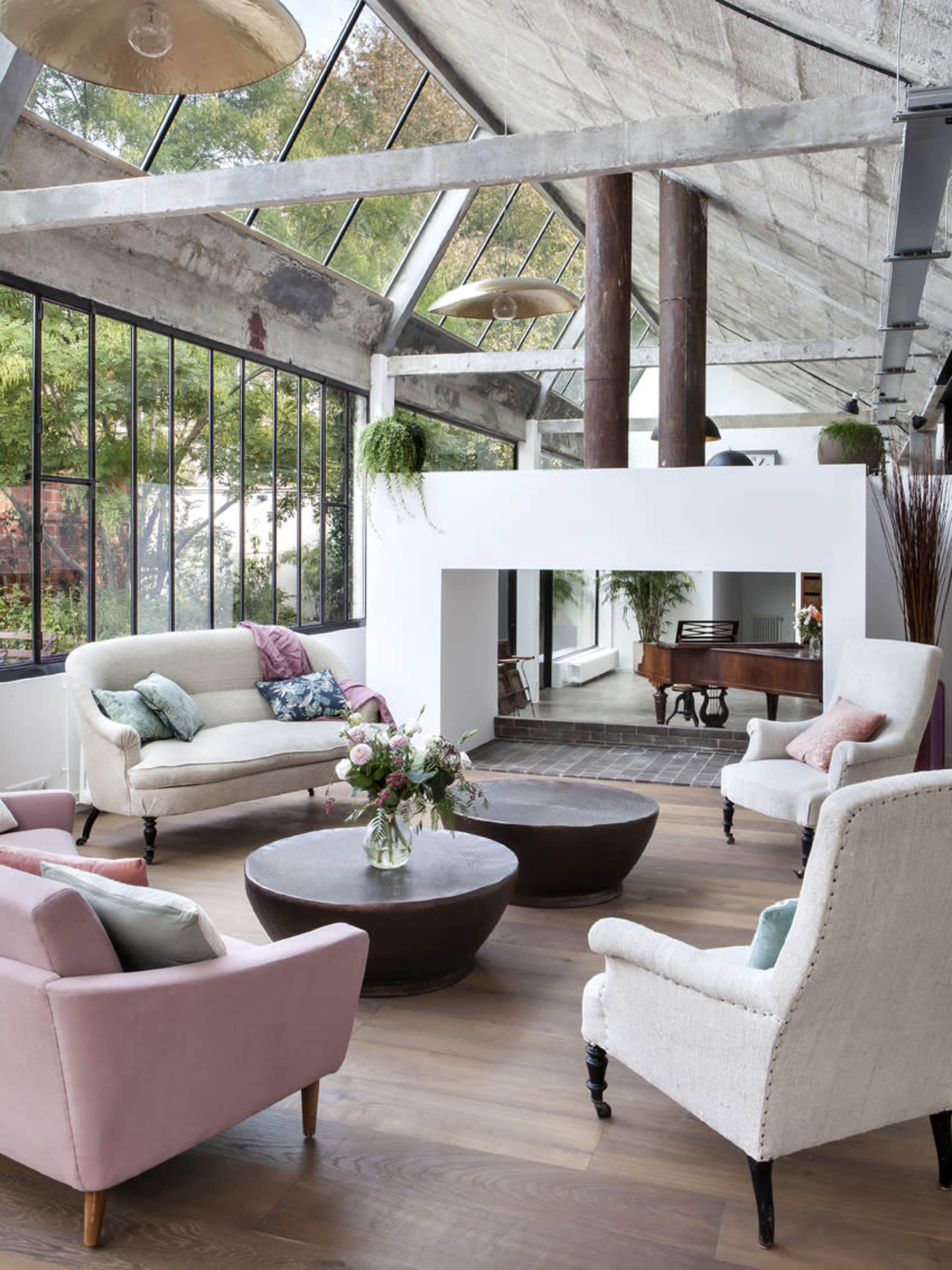 The image shows a modern living room with large windows, featuring a round coffee table surrounded by several armchairs and a white fireplace.