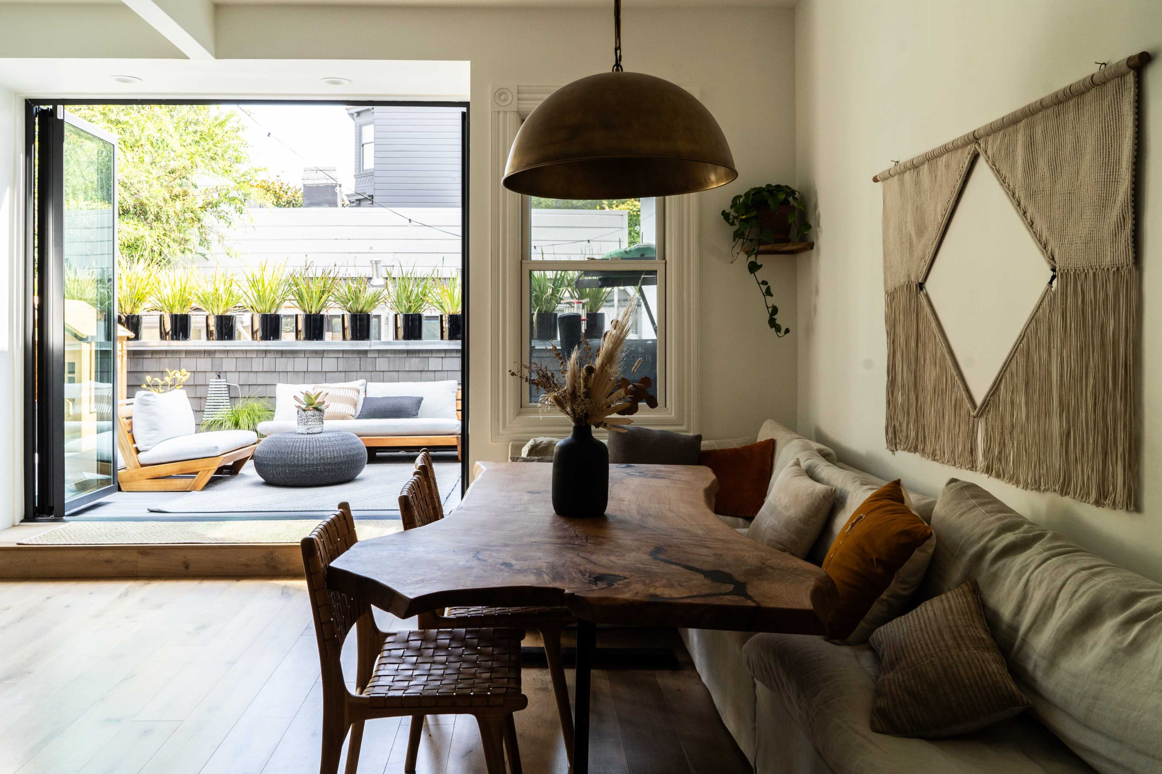 A modern living space features a large wooden table surrounded by chairs, with an open door leading to a patio filled with greenery and comfortable outdoor furniture.
