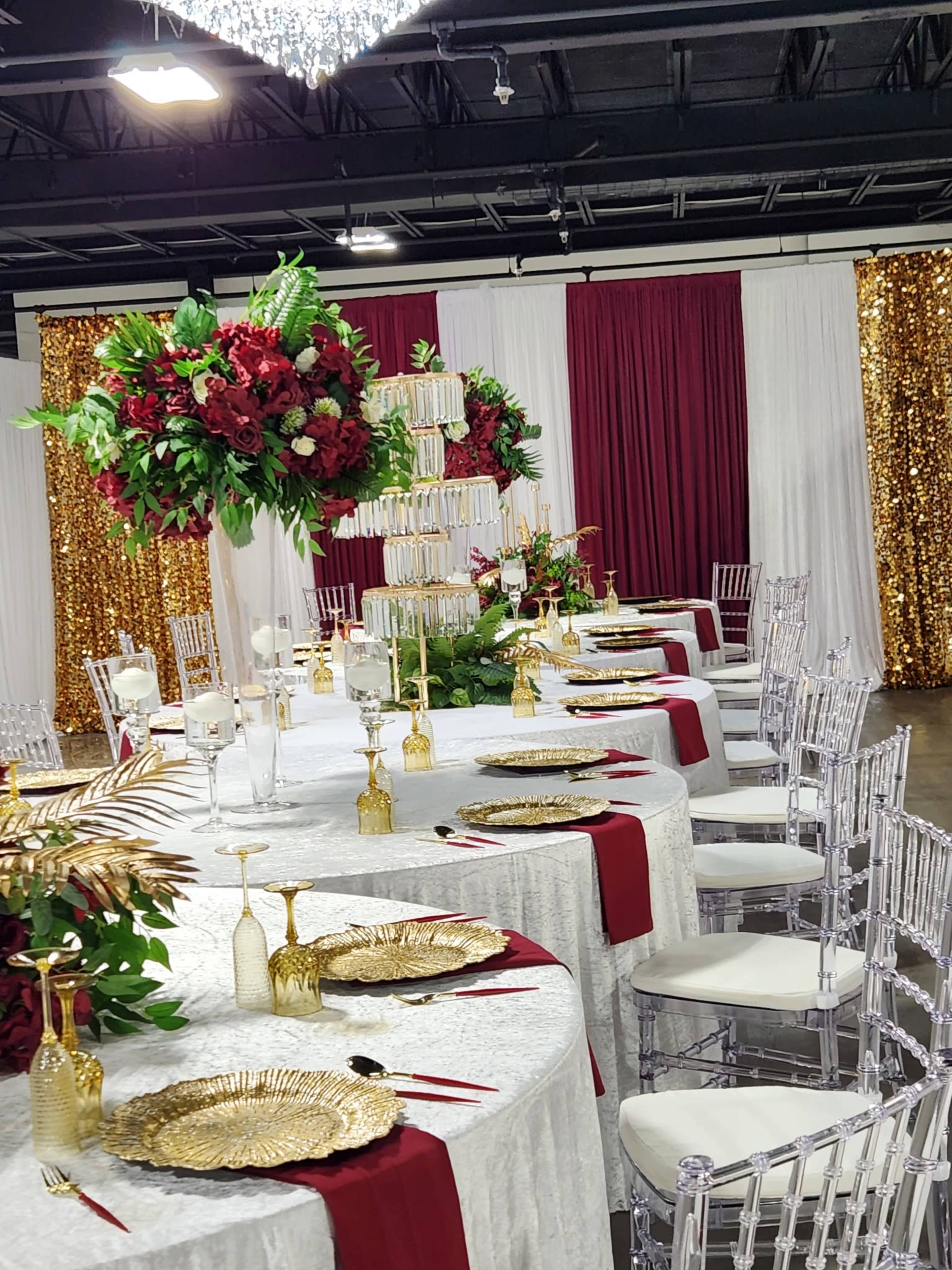 The image shows an elegant banquet setup with tables draped in white and red fabrics, adorned with floral centerpieces and gold accents, and surrounded by clear acrylic chairs.