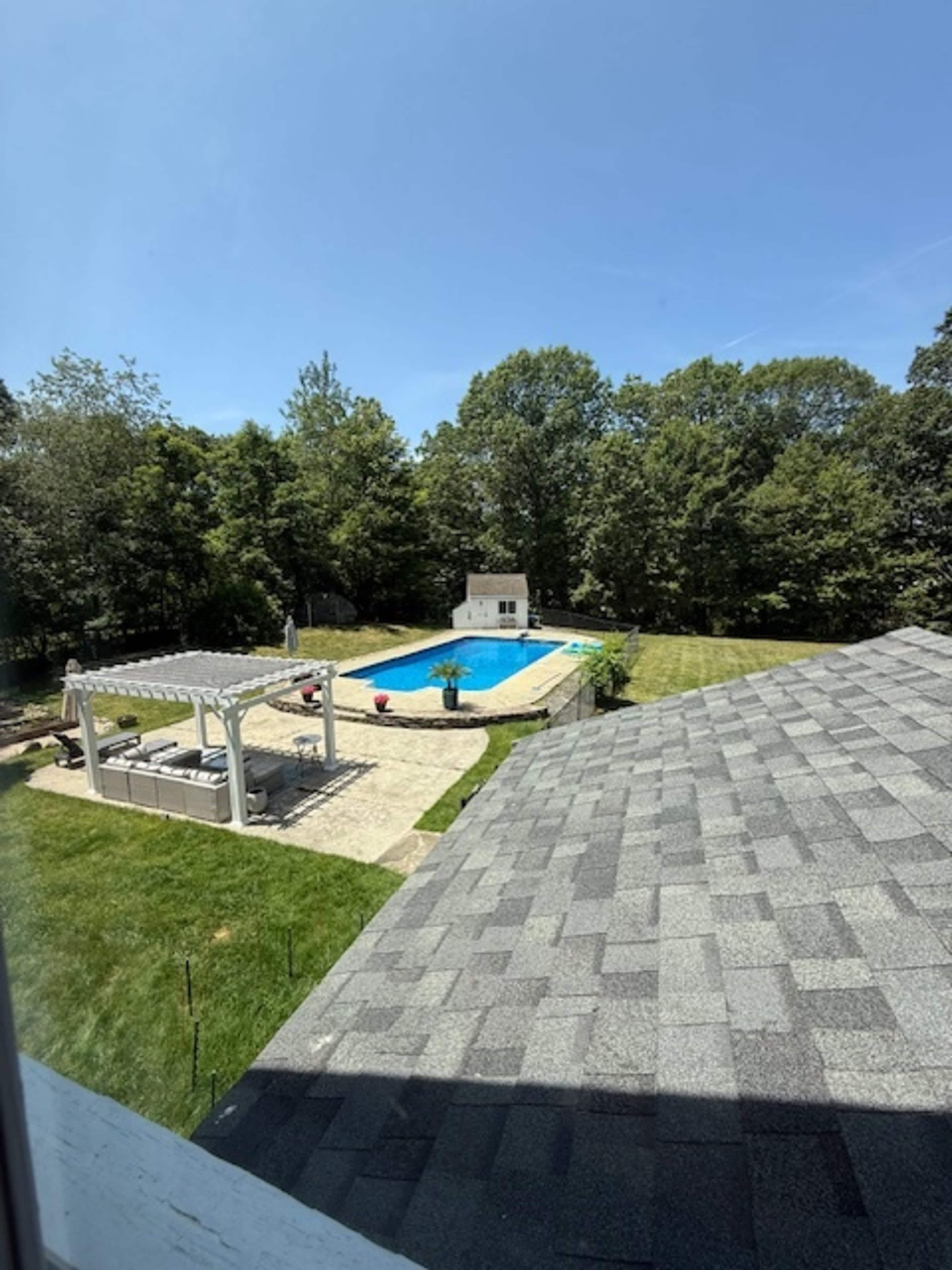 The image shows a backyard with a swimming pool, surrounded by a grassy area and trees, with a pergola and seating arrangement nearby.