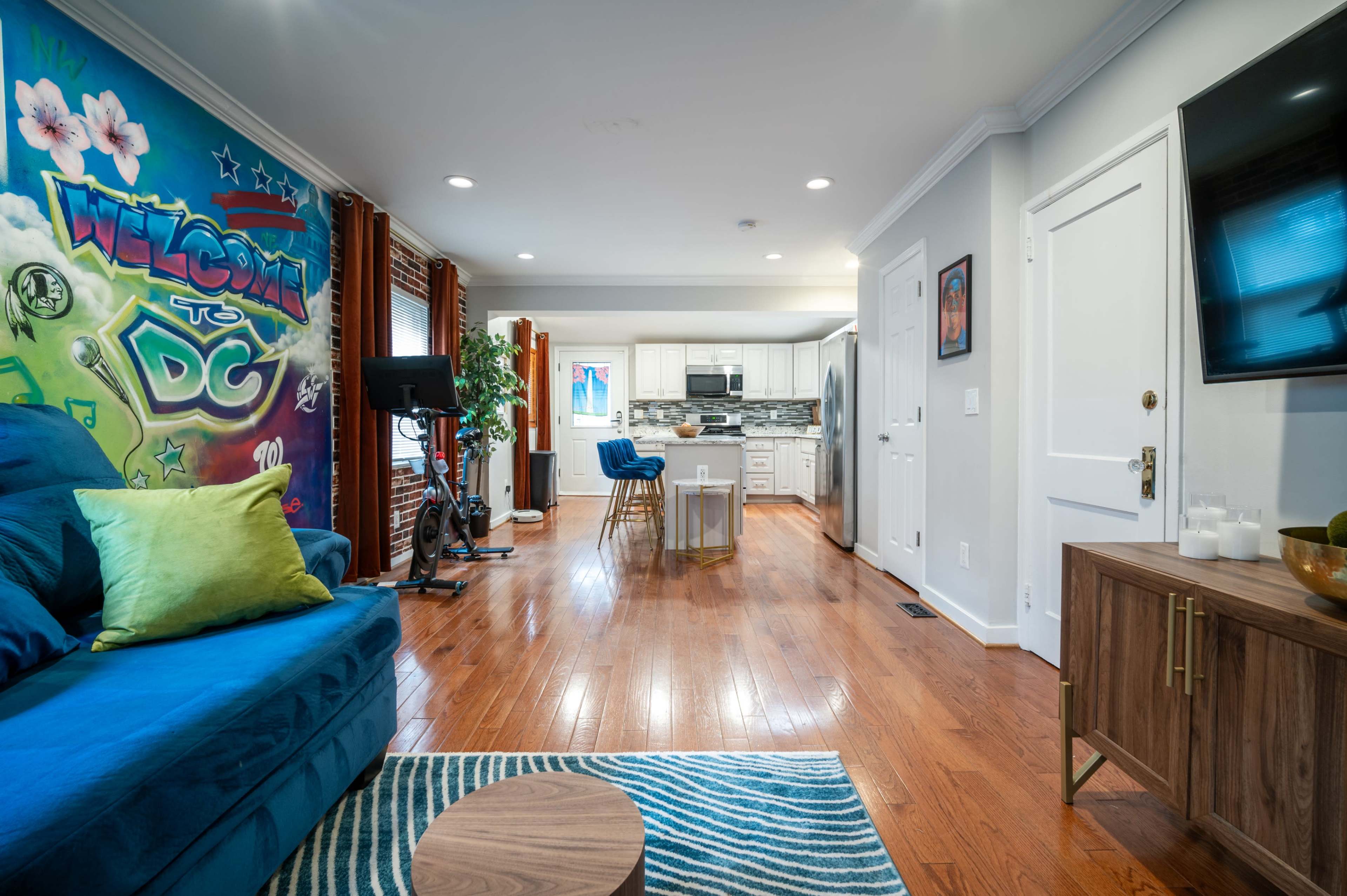 A brightly painted living area features a blue couch, a wooden coffee table, and a view of a kitchen with white cabinets and modern appliances.
