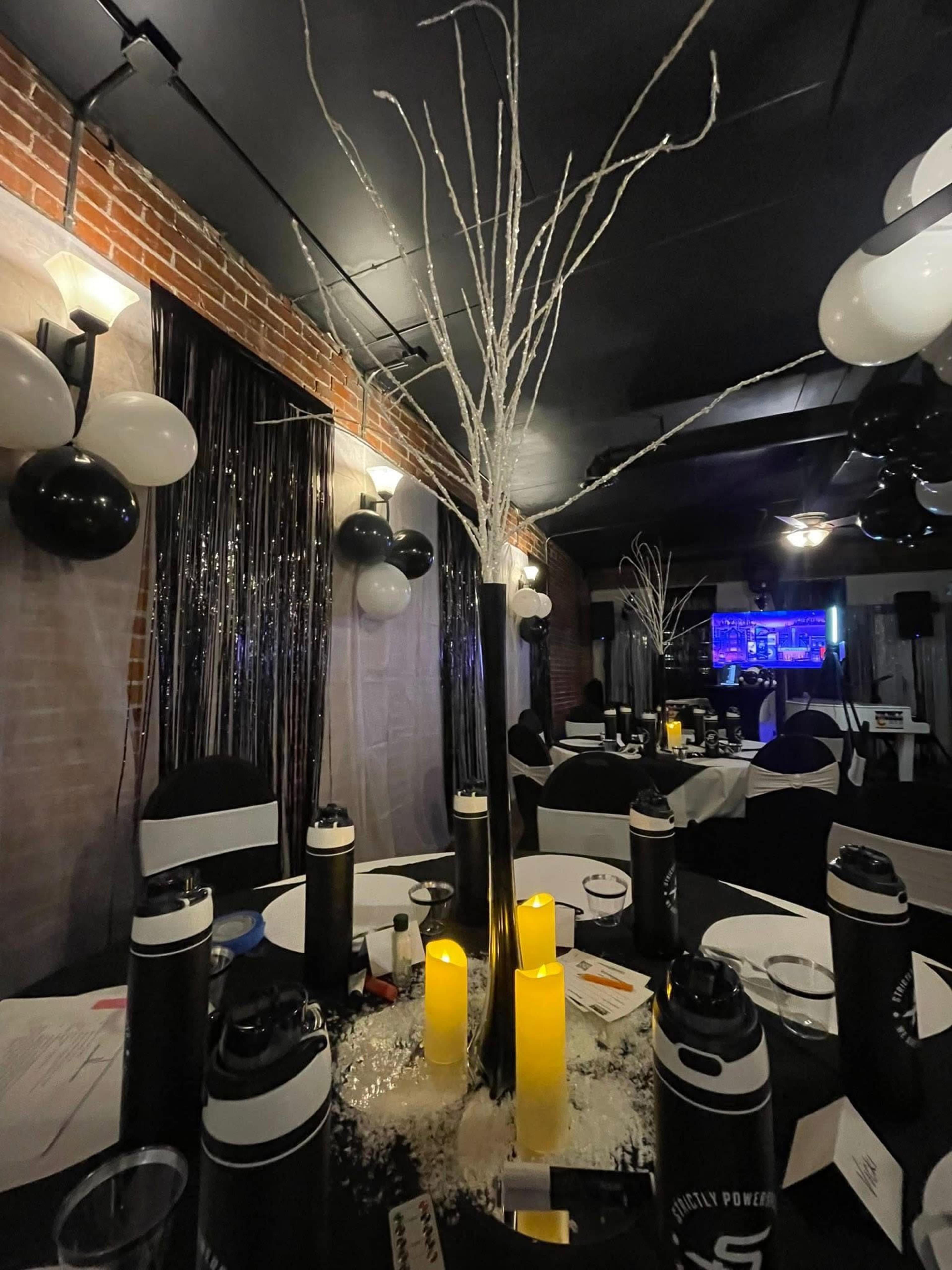 A banquet table is decorated with white branches, candles, and black-and-white balloons in a dimly lit venue.