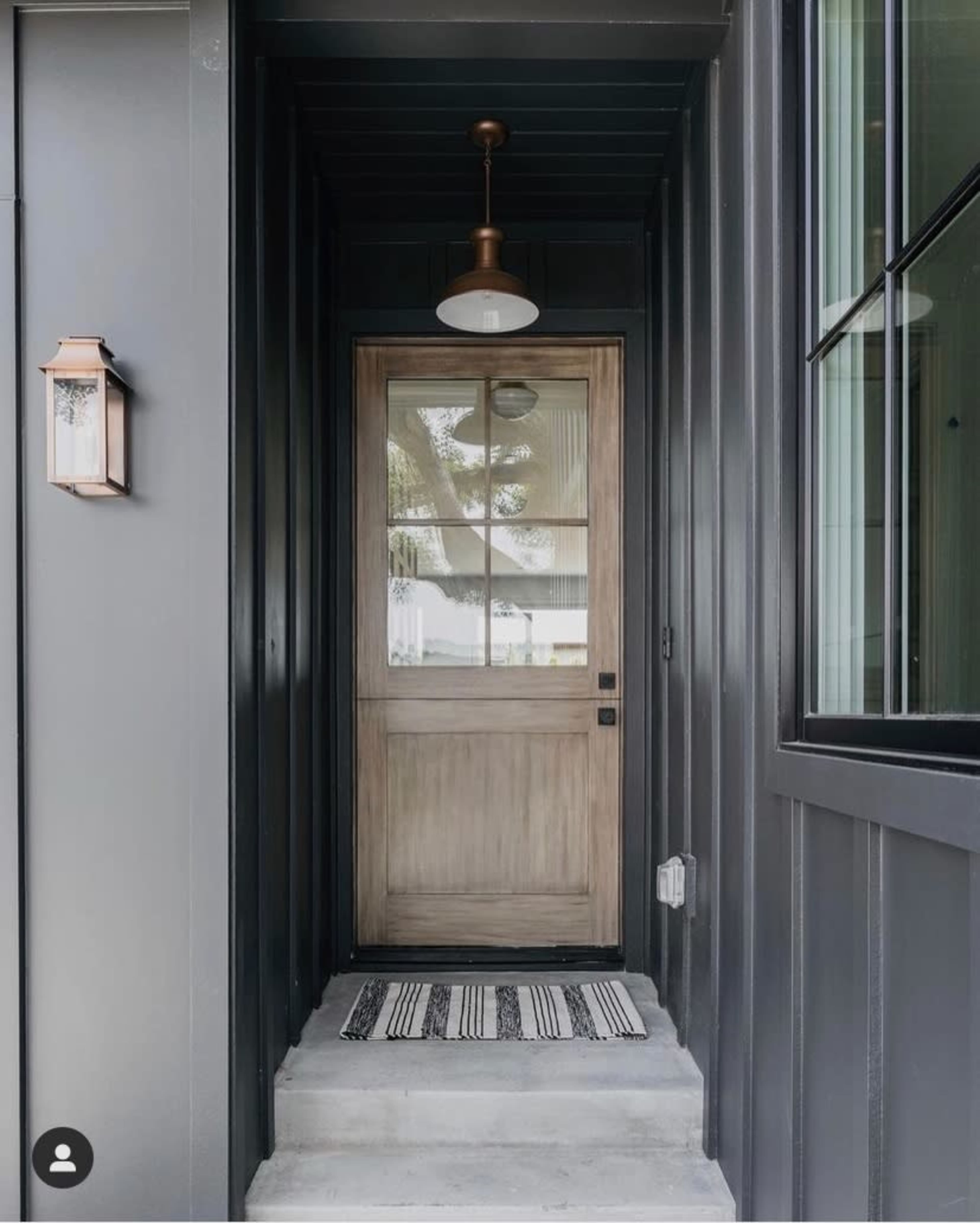 The image shows a modern entrance with a wooden door, a light fixture above, and a black and white striped doormat.