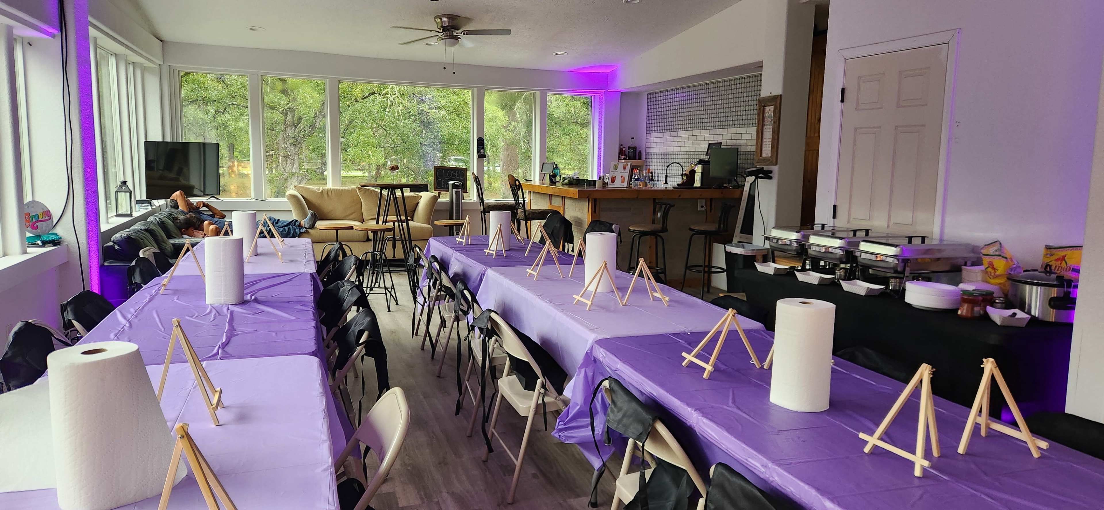 The image shows a spacious indoor area set up with several rows of purple-covered tables and easels, equipped for an art activity, alongside a bar and a seating area.
